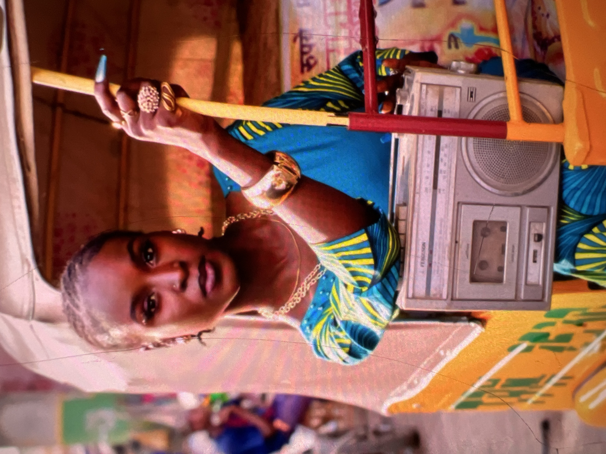 A medium shot captures a woman with braided hair, adorned with earrings, a gold chain, and several rings on her hand, which grips a yellow pole. She wears a blue garment with yellow and green stripes. A silver boombox sits on a colorful structure beside her. The background shows a blurred street scene, suggesting an urban environment, likely Lagos, Nigeria, given the presence of what appears to be an auto-rickshaw or "keke napep." The overall impression is one of vibrant street life, with the woman and the boombox as focal points.