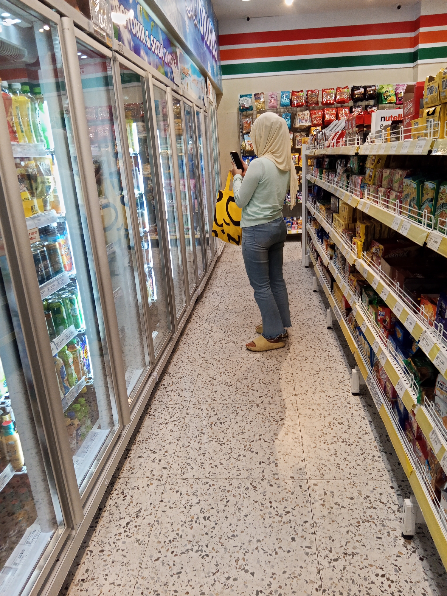 A woman wearing a hijab and light blue jeans stands in an aisle of a 7-Eleven convenience store, holding a smartphone and a yellow bag. She is facing away from the camera, looking at the shelves filled with various snacks and products. The store features large refrigerated display cases filled with drinks on the left side of the image and brightly colored shelves stocked with merchandise on the right. The floor is tiled with a speckled pattern. The store is likely located in Lubuklinggau, Indonesia, given the context provided.