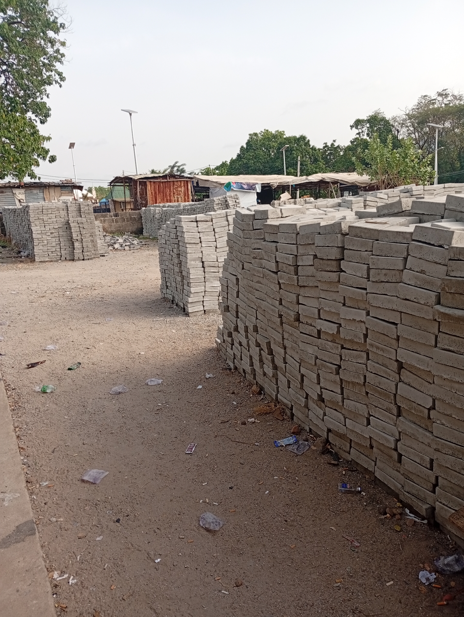The image depicts a dusty, unpaved area in Adankolo, Nigeria. Several stacks of rectangular, gray concrete blocks are arranged in the foreground and midground. These blocks appear to be construction materials or paving stones. The ground is littered with various small pieces of debris, including plastic wrappers and other unidentified objects. In the background, there are simple, partially dilapidated structures, possibly market stalls or sheds, with metal roofs and wooden supports. Tall light poles are also visible in the distance. The scene lacks any visible human activity. The overall impression is one of a storage area for construction materials in a somewhat impoverished or underdeveloped urban environment.