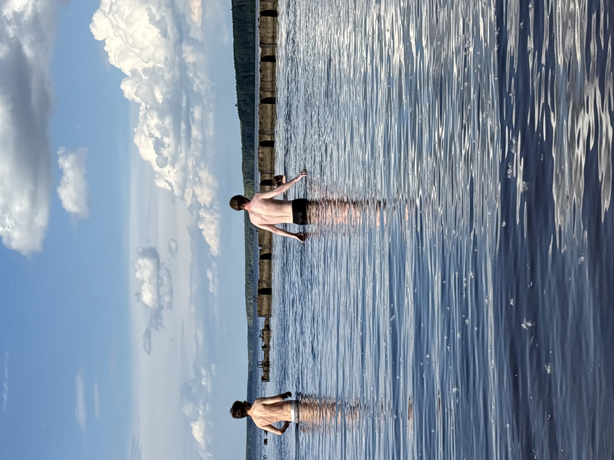 The image shows two young men in the water. One man, in the foreground, is partially submerged from the waist down, with his back to the camera. The second man is further in the background, also partially submerged from the waist down, facing away from the camera with his arms outstretched. Both appear to be enjoying the water. The setting is a body of water, possibly a lake or river, with a shoreline in the distance that features a dense line of trees. A line of old, weathered pilings or posts runs horizontally through the water. The sky is blue with fluffy white clouds, suggesting fair weather and daytime. The water is calm with gentle ripples reflecting the sky and the surrounding environment. There is no visible text in the image. The context provided indicates the location is Chaikovsky, Russia.