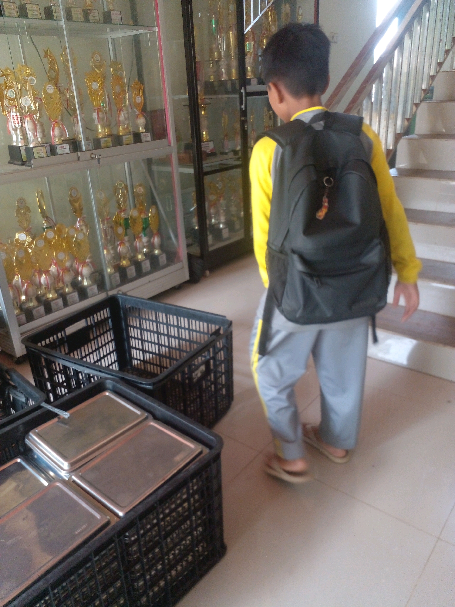 A young boy wearing a yellow long-sleeved shirt, grey pants with a yellow stripe, and a black backpack stands in what appears to be a trophy display room. To his left are two black plastic baskets, one of which contains several metallic trays. In the background, glass display cases filled with gold trophies are visible. The floor is tiled and there is a staircase to the right of the boy. The boy is facing away from the camera and appears to be walking. The scene suggests a school or an institution where awards are given.