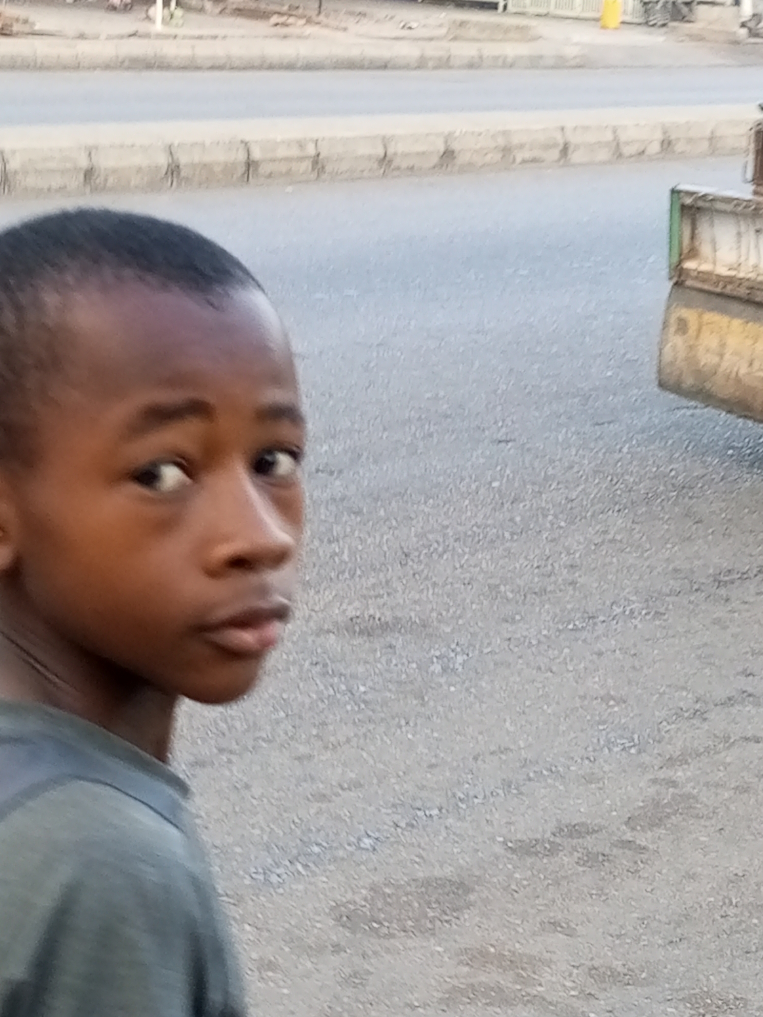 A close-up, eye-level shot captures a young boy looking directly at the camera, with a slight turn of his head and body to his right. The boy's face is the primary focus, with his gaze directed slightly to his left, away from the viewer. He is wearing a grey t-shirt. The background is blurred, but appears to be an outdoor urban environment. A concrete curb and a paved road are visible. To the right of the frame, a portion of a vehicle, possibly a truck bed with green and yellow accents, is visible. The lighting suggests it is daytime, with the sun casting no harsh shadows. The overall impression is one of a candid, street-level portrait.