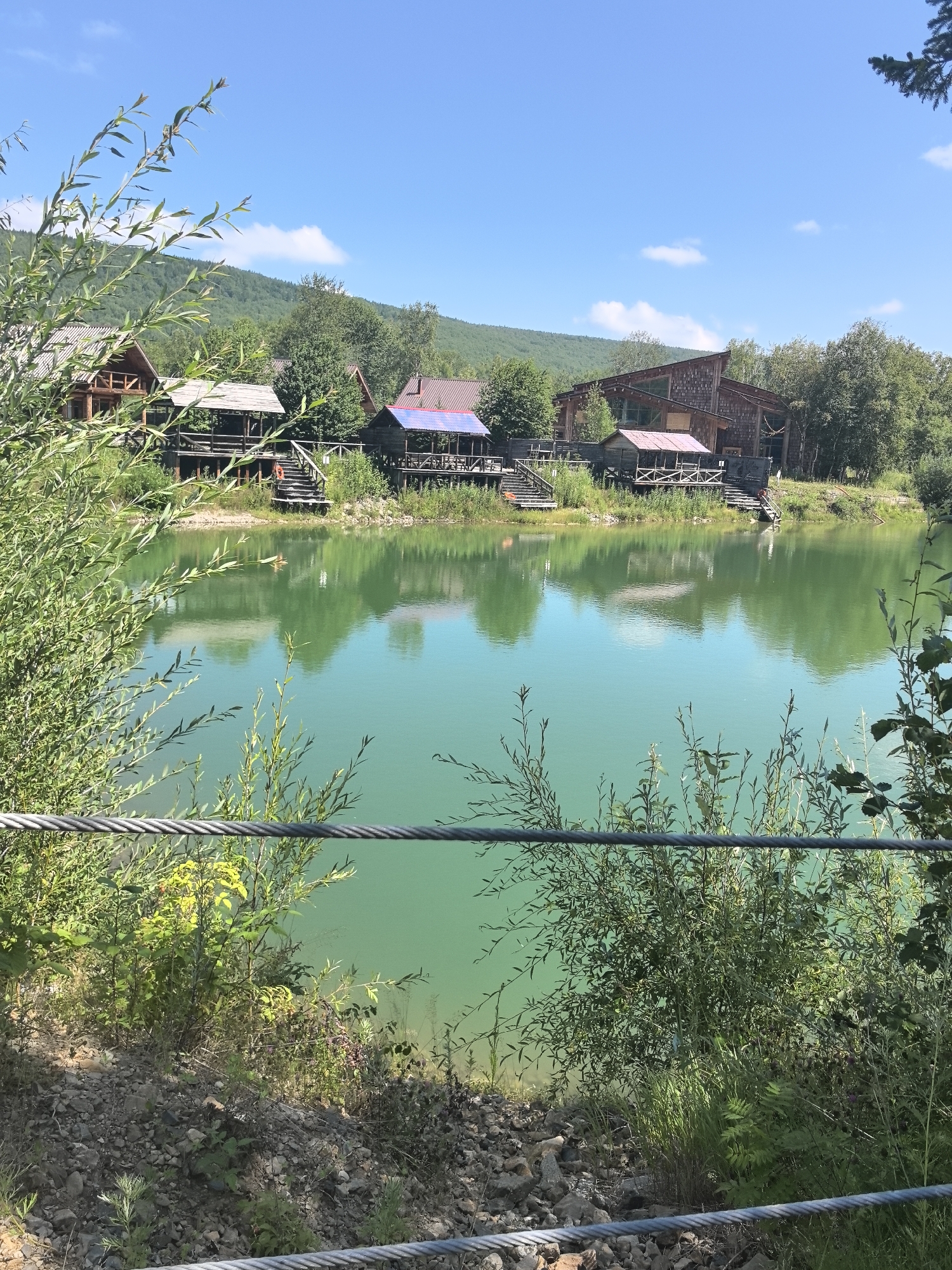 A scenic view of a tranquil turquoise lake is captured on a clear, sunny day. Several wooden structures, resembling cabins or lodges, are situated on the opposite bank, nestled among lush green trees and rolling hills. These structures feature wooden decks and some have a rustic, natural aesthetic, with one appearing to have a blue roof. The calm water perfectly reflects the sky, clouds, and the surrounding vegetation, creating a serene and picturesque landscape. In the foreground, thick green foliage and branches partially obscure the view, with a thick metal cable running horizontally across the lower portion of the frame, suggesting a barrier or a railing. The ground in the immediate foreground is rocky and covered with scattered stones and low-lying green plants. The overall impression is one of a peaceful natural retreat. The location is in Solnechnyi, Russia.