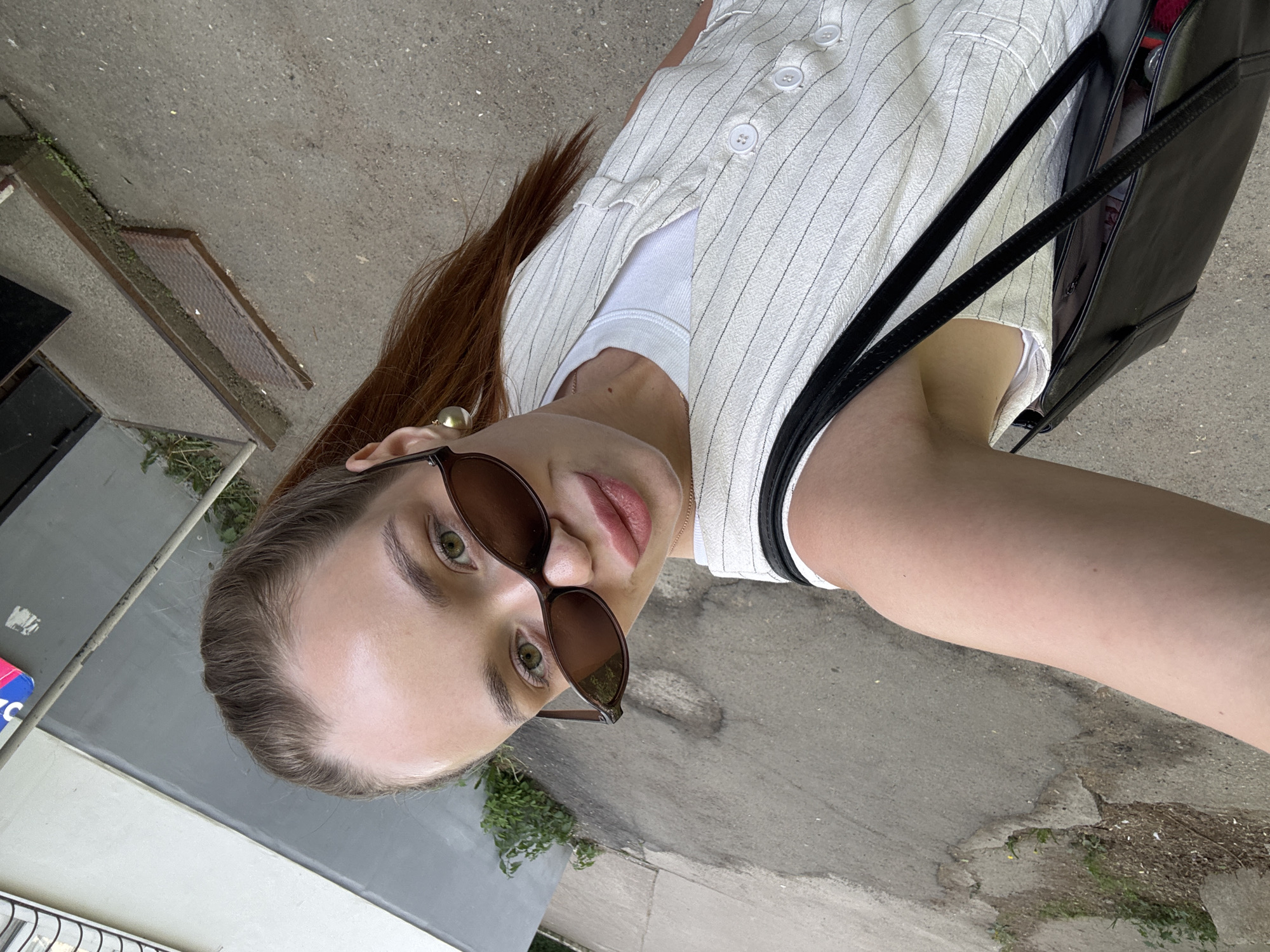 A young woman with reddish-brown hair is wearing sunglasses, a white t-shirt, and a pinstripe button-down shirt. She has a black purse or bag on her shoulder. The background shows a gray concrete surface, possibly a sidewalk or street. There is some greenery growing in cracks in the concrete. It appears to be an outdoor setting. The location is Chaikovsky, Russia.