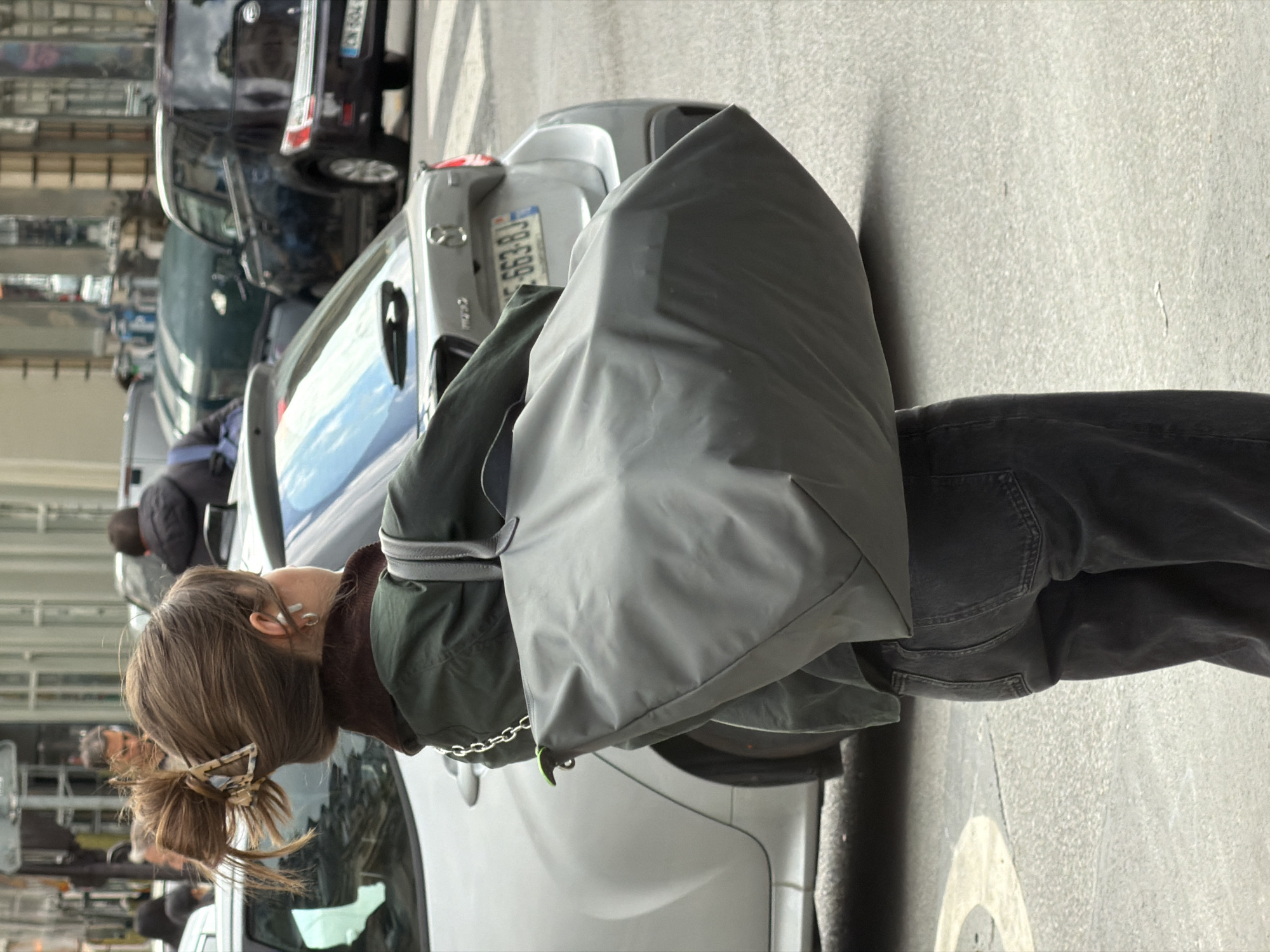 A street scene in Paris, France, is captured in a vertical, medium shot. A woman stands near a parked, silver-colored Mazda. She wears dark jeans and a green jacket. A large gray bag is slung over her shoulder. Her hair is pulled back with a tortoiseshell clip. In the background, a dark-colored vehicle is parked. The location is identifiable as Paris, France.