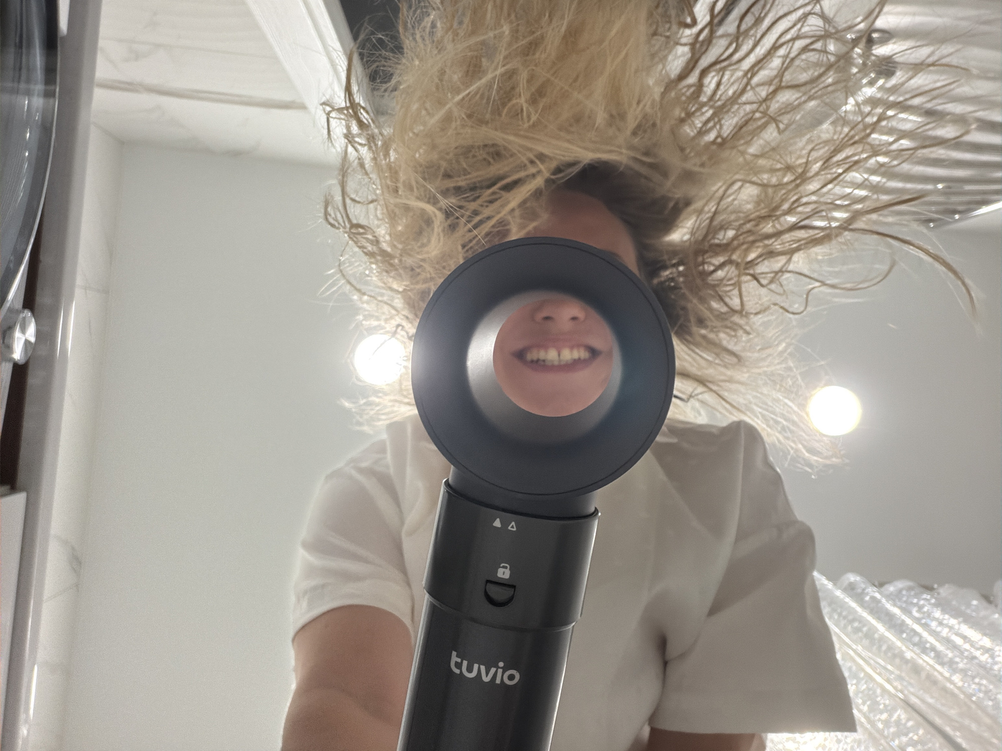 A low-angle shot captures a person holding a "tuvio" brand hair dryer up towards the camera, with the dryer's nozzle framing their mouth in the center. The person has blonde, tousled hair filling the upper portion of the frame. They wear a white t-shirt. The background is a bright, sterile looking bathroom with white surfaces and light fixtures. The location in Votkinsk, Russia is not discernible from this image.