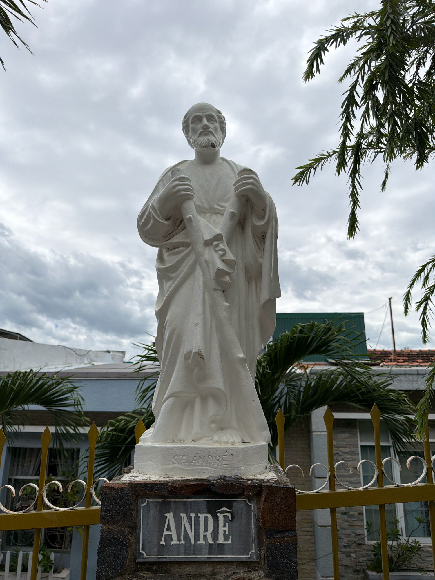 The image captures a white marble statue of a bearded man, presumably Saint Andrew, standing on a stone pedestal. The statue is dressed in a flowing robe and holds a staff. Inscribed on the pedestal is "ST. ANDRE," and below that, on a dark plaque, the name "ANRE" is visible. The statue is positioned in front of a yellow wrought-iron fence. Behind the fence, there are palm trees and a building with light blue and grey stone walls. The sky is overcast with clouds. This statue is located at the intersection of Quang Trung and Tran Phu streets in Nha Trang, Vietnam, near the Nha Trang Cathedral.