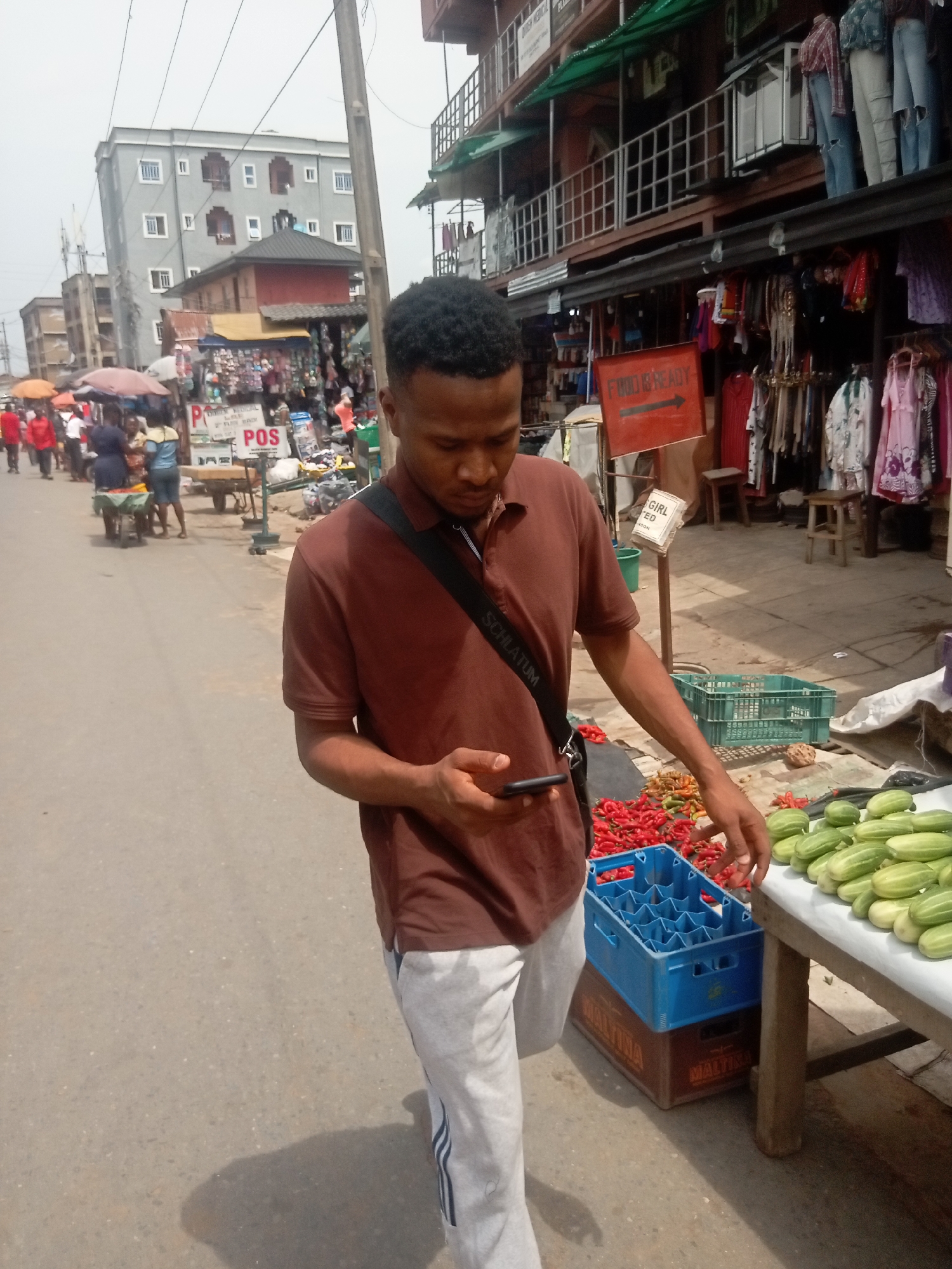 A man in a brown polo shirt and gray sweatpants stands on a road next to a table piled with cucumbers and red peppers. He is holding a mobile phone. A blue plastic crate sits atop a brown cardboard box on the ground near the table. Behind him is a bustling street scene with vendors and pedestrians. Buildings line the street, some with clothes and other goods displayed for sale. A sign pointing towards "Food is ready" is also visible. The location is Onitsha, Nigeria, based on the maltina brand on the cardboard box.