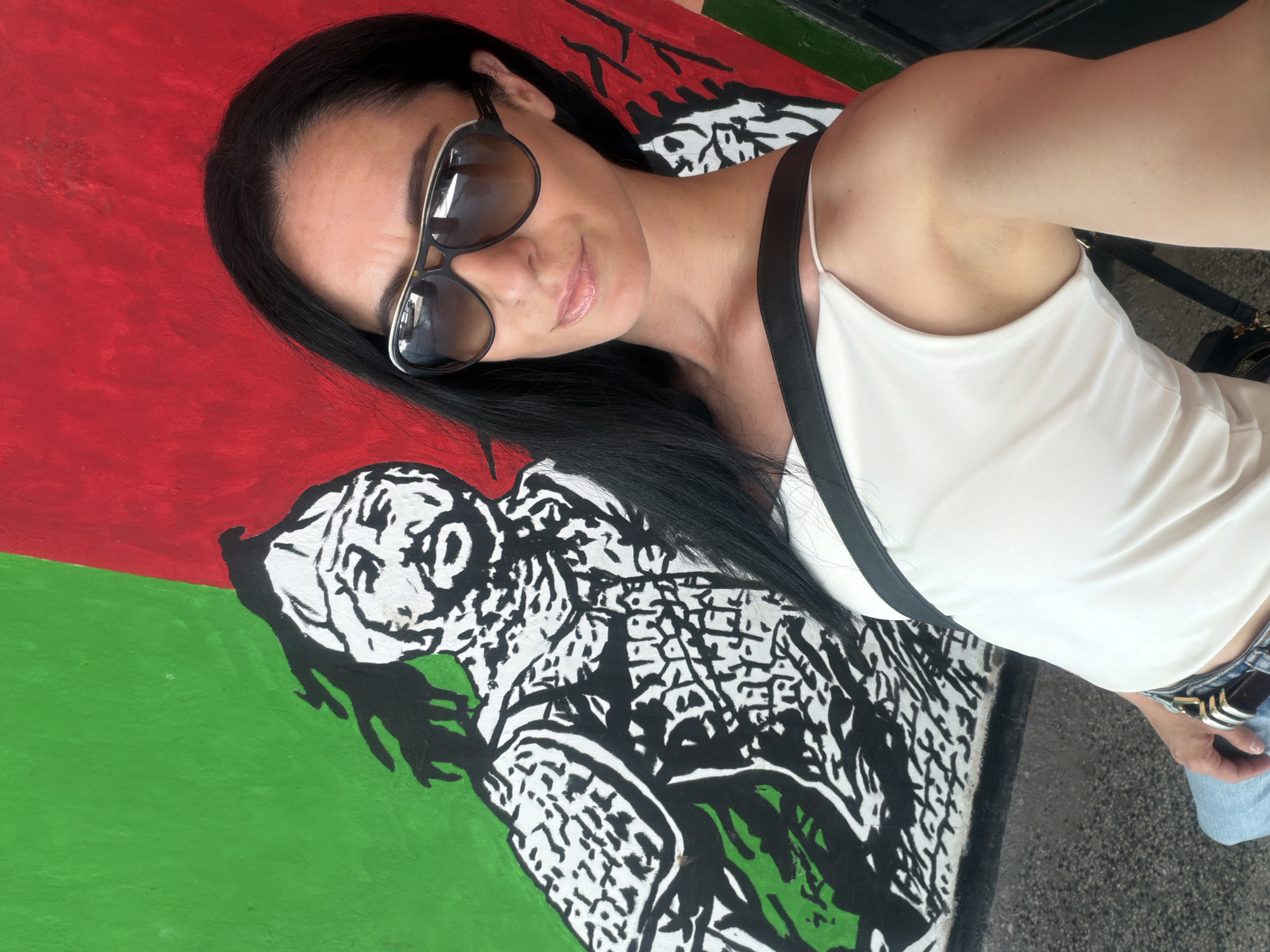 A selfie captures a woman posing outdoors in front of a colorful mural in Pleven, Bulgaria. The woman, with long, straight dark hair, is seen from the chest up, angled diagonally. She wears stylish large, dark sunglasses with silver accents, which obscure her eyes, and a light-colored (possibly white or cream) spaghetti-strap top. A dark strap, likely from a bag or part of her clothing, crosses her left shoulder. A slight, closed-lip smile suggests a pleasant and relaxed demeanor. Her lower attire, visible at the bottom right, includes blue jeans and a brown belt with a silver buckle.
The background is a vibrant mural featuring large, distinct sections of solid red and green. Over these colored areas, intricate black and white graphic designs are painted. The red section displays abstract, angular black and white patterns, while the green section showcases a stylized depiction, possibly of a creature's head or a human-like face, rendered in bold black and white lines. The ground beneath the mural and the woman appears to be dark asphalt or concrete. The bright, clear lighting indicates that the photo was taken during the daytime in sunny weather. There is no visible text in the image.