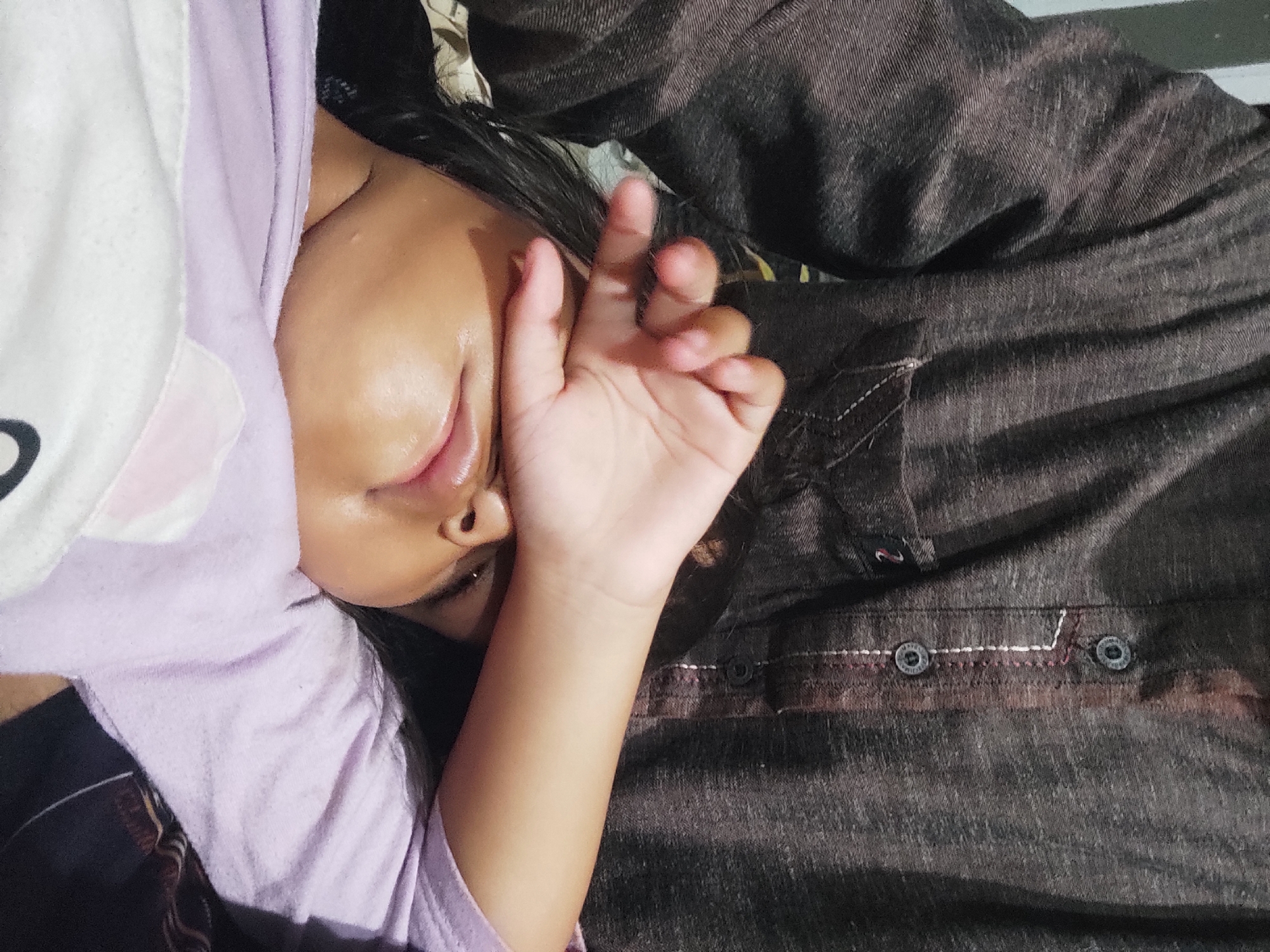 In Sumedang, Indonesia, a close-up shot shows a young child resting their head on a person's chest, with one hand covering their eyes. The child wears a light purple shirt. The adult, partially visible, is dressed in a dark brown textured shirt with buttons and decorative stitching. The background is indistinct.