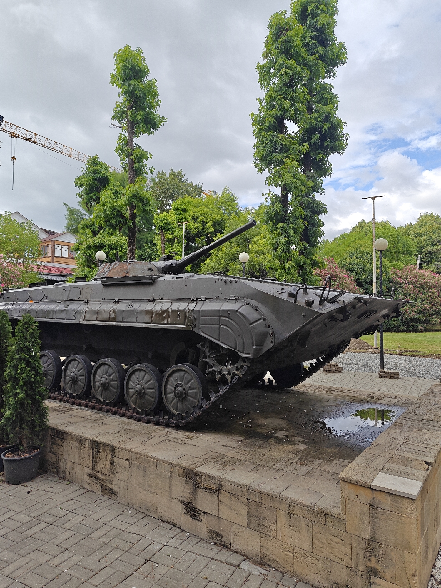 An olive-drab tank is displayed on a stone pedestal outdoors. The tank is viewed from the right side, with its tracks visible. Several large road wheels are on the right side of the tank. The tank is positioned on a paved area with a brick border and a small puddle of water. Behind the tank are several trees, with one tall, narrow tree directly behind it. In the background, there are buildings and a construction crane. The sky is overcast with some patches of blue.
