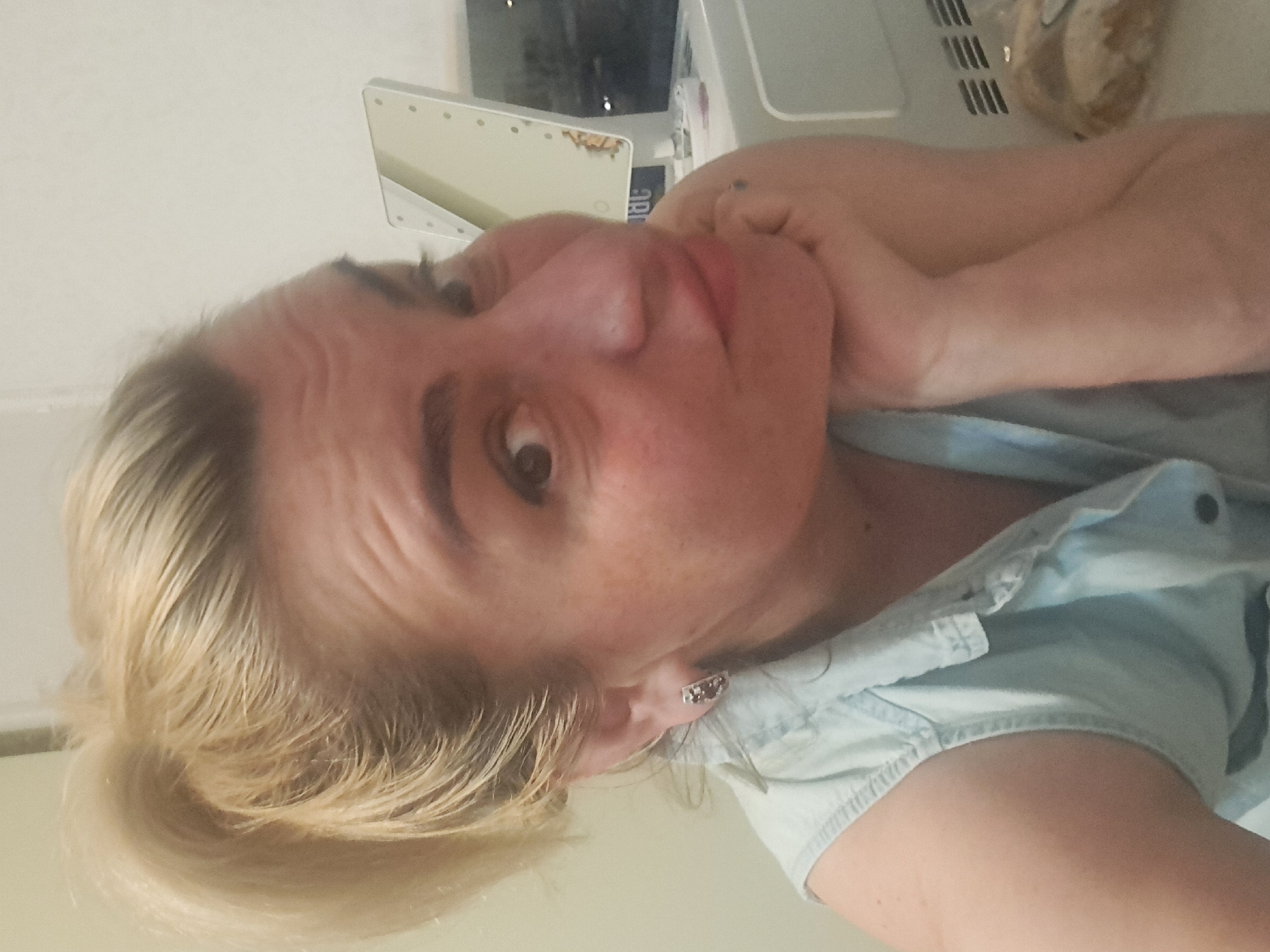 A woman with blonde hair and fair skin is posed in a selfie. She is wearing a light blue denim shirt and has earrings. Her face is partially supported by her hand. Behind her are a lighted vanity mirror and other objects on a countertop. The photo appears to be taken indoors, possibly in a bathroom or bedroom. There is no information that this photo was taken in Elektrostal, Russia.