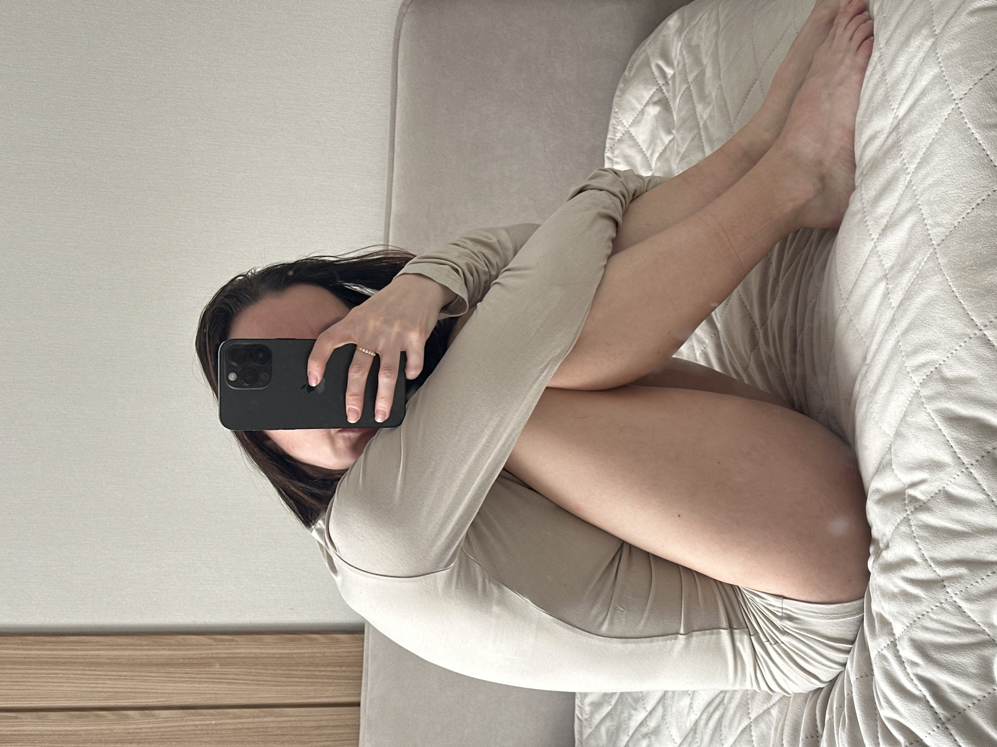An individual is seated in a fetal-like position on a bed within an indoor setting in Votkinsk, Russia. The person, appearing female, wears a light beige long-sleeved bodysuit. Her dark hair is visible around her head and shoulders. She holds a dark-colored smartphone, featuring a multi-lens rear camera system, vertically in her left hand, positioning it to obscure her face. A ring with small, clear stones is present on her left ring finger. Her bare legs are drawn towards her torso, with her arms wrapped around them. The bed is covered with a light beige quilted duvet displaying a diamond pattern. Behind the individual is a soft, upholstered headboard or backrest of a similar light beige color. The background consists of a plain light-colored wall, with a section of vertical light-toned wooden paneling visible on the left side of the frame. The scene is illuminated by soft, ambient light.