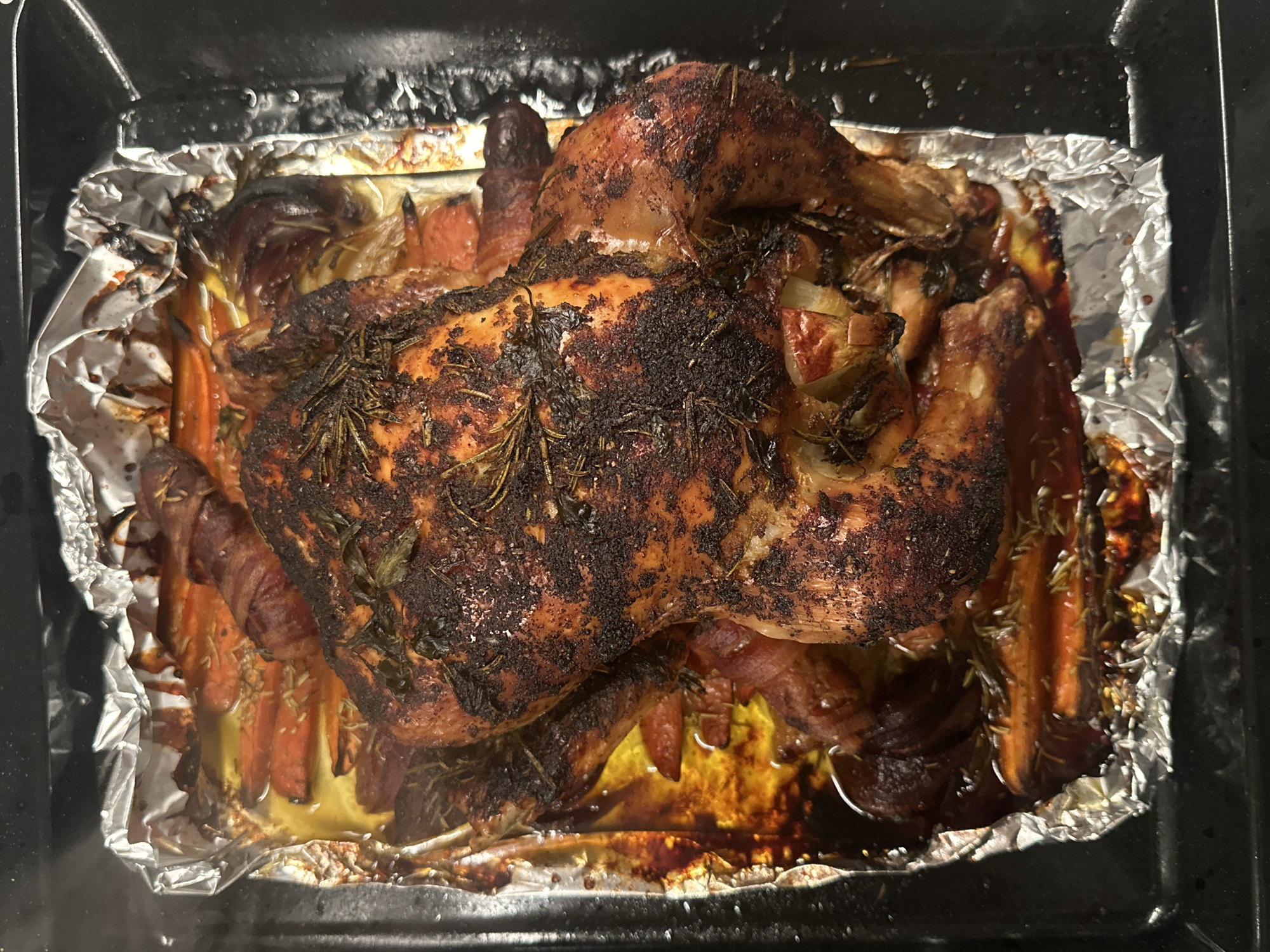 A roasted chicken rests atop sliced carrots, onions, and possibly other root vegetables in a foil-lined baking pan. The chicken is browned, possibly over-cooked, with herbs sprinkled on top. The baking pan is inside a black oven. No location information or details about people or interactions are discernible from the image.