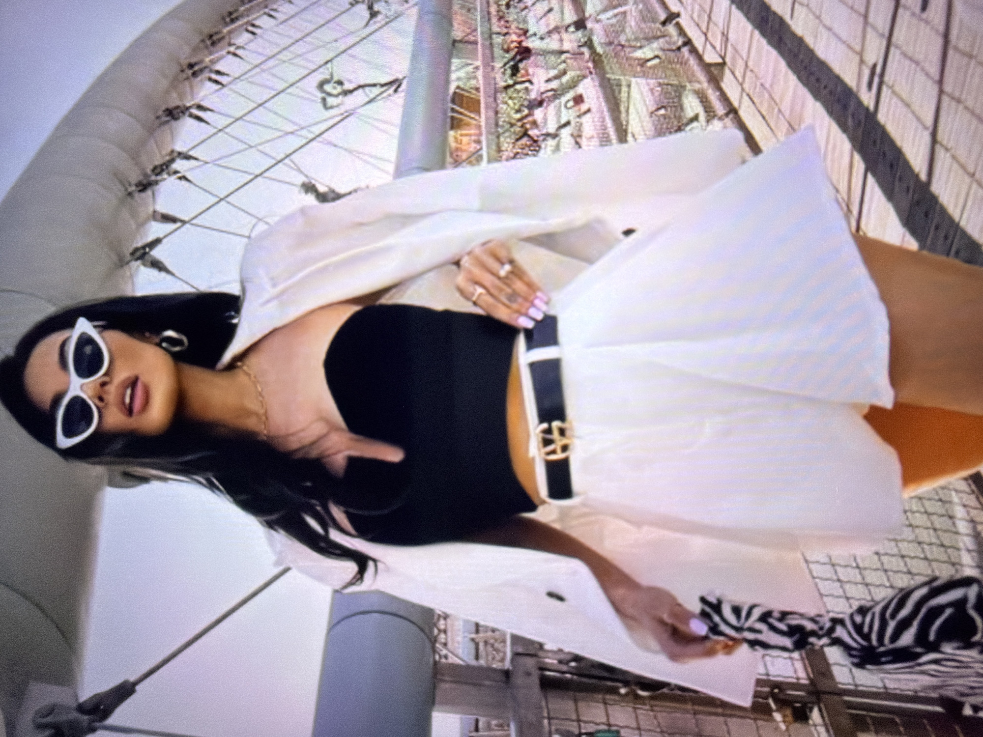 A woman stands on a pedestrian bridge, posing with a zebra-print bag. She is wearing a black strapless top, white pleated shorts, a white blazer, a black belt with a gold buckle, and white cat-eye sunglasses. The bridge has a metal railing and the walkway is made of wooden planks. In the background, there are numerous locks attached to the railing, suggesting it's a "love lock" bridge. The sky is bright and overcast. The location is identified as the city of Beșalma, Moldova.