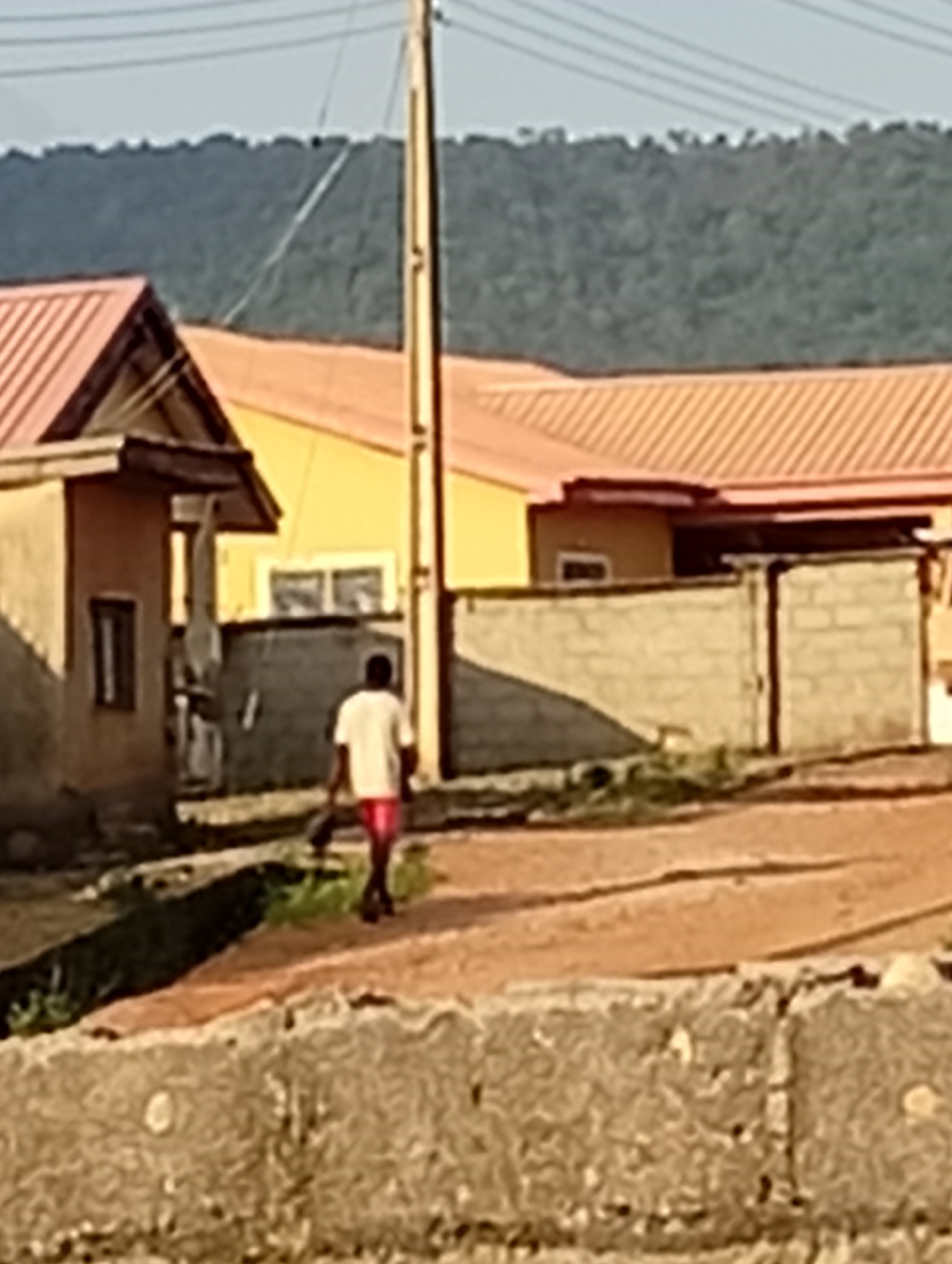 A residential area in an unknown city features a man walking away from the camera on a dirt road. He is wearing a white t-shirt and red shorts and carrying something dark in his left hand. Several buildings with red-tiled roofs are visible, along with a cinder block wall and a utility pole. A forested hill is in the background under a bright sky.