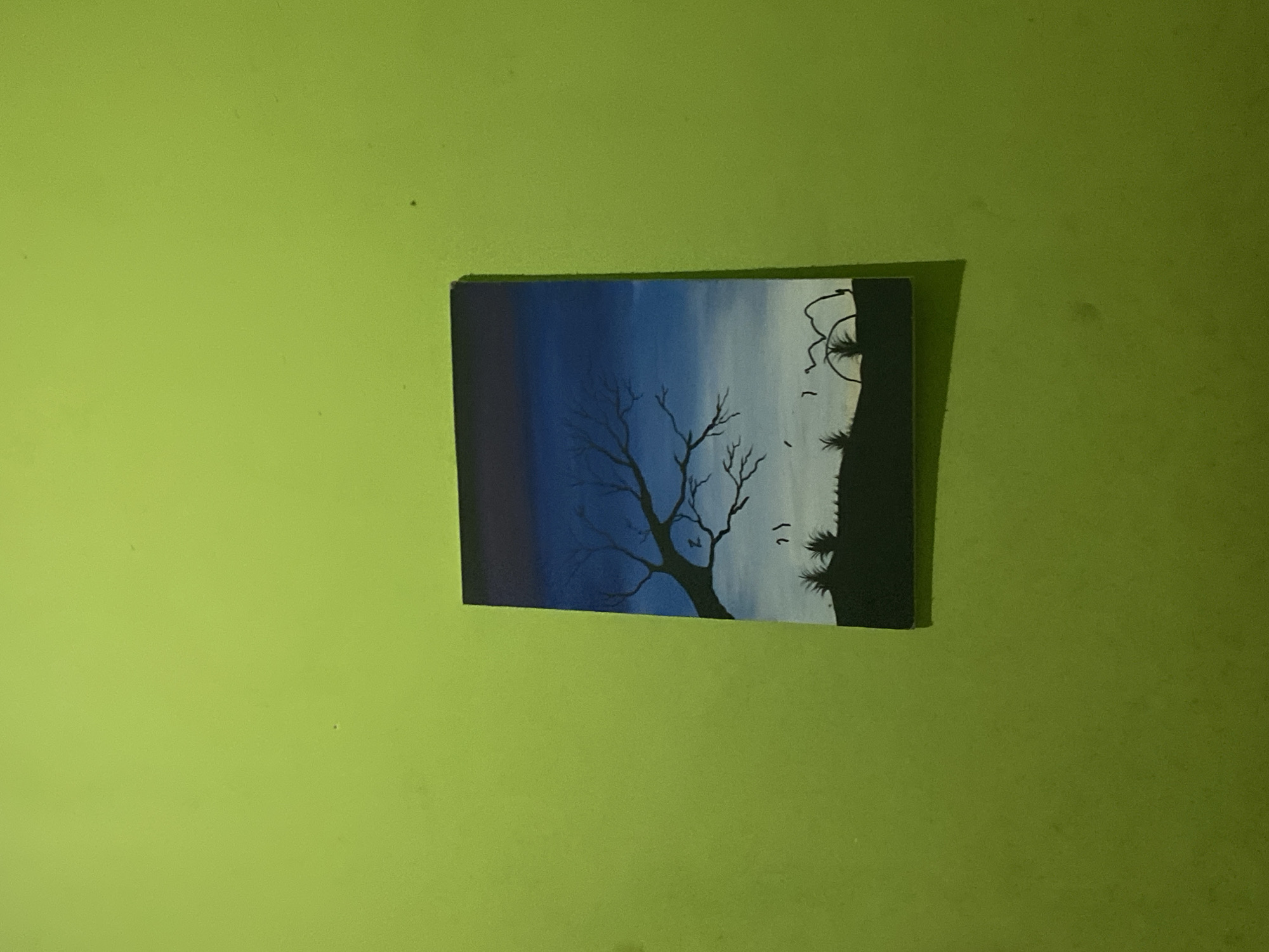 A painting is hanging on a bright green wall. The painting depicts a silhouetted landscape with a bare tree in the foreground. The sky transitions from dark purple at the top to blue and then white near the horizon. There are small bird-like shapes in the sky and some tufts of grass or bushes on the ground. The painting is rectangular and appears to be hung slightly askew. The green wall is textured and shows some minor imperfections. The setting is indoors, with no indication of the city of Ta', Indonesia.
