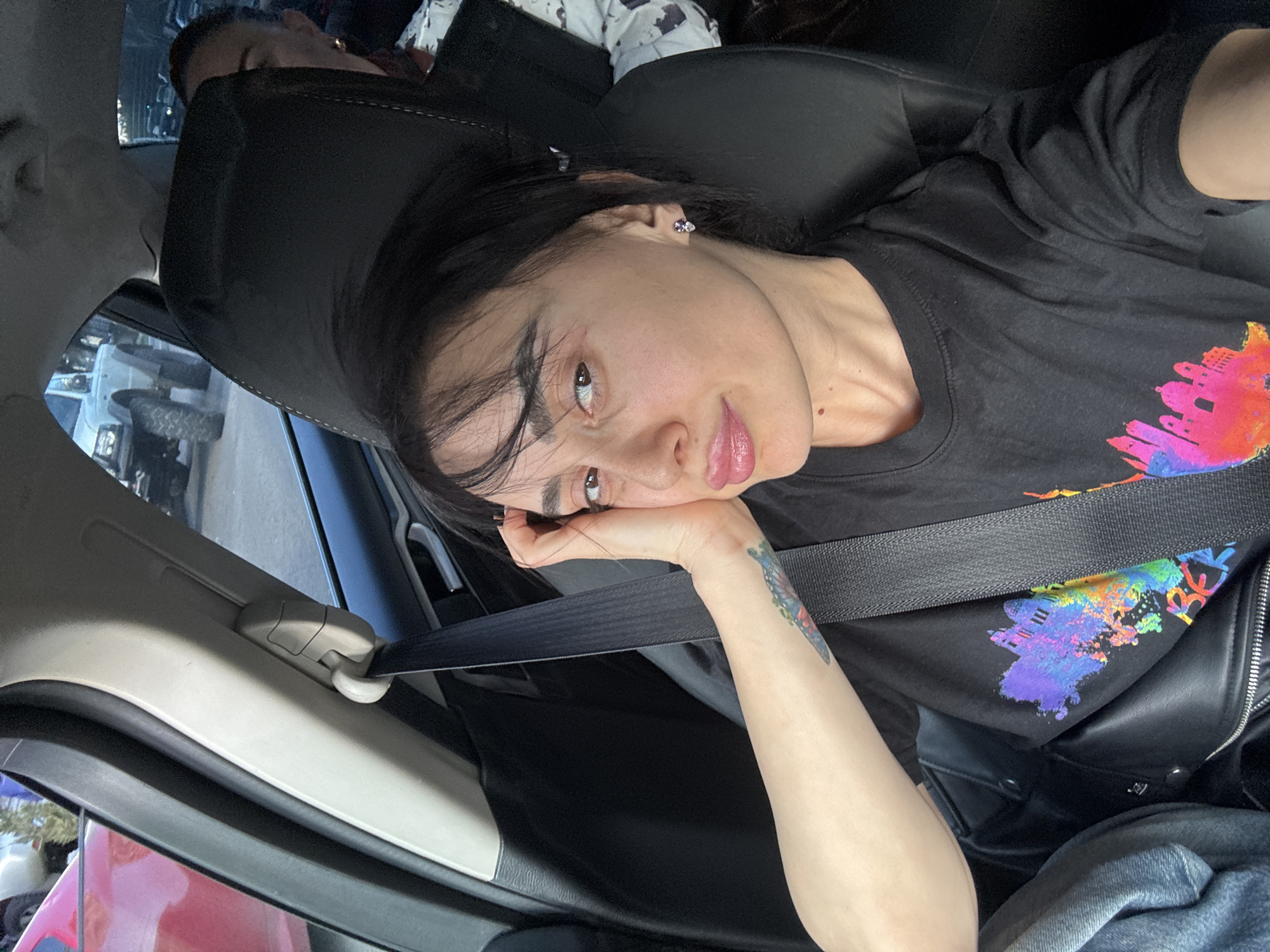 A woman with dark hair is sitting in a car wearing a black T-shirt with a colorful building design on the front, a black leather jacket, and a seatbelt. She has a tattoo on her arm. She is looking towards the camera. Another person is visible in the back seat. Outside the car, there are other cars, suggesting a city street. No specific building or feature is visible to pinpoint the exact location in Tbilisi, Georgia.