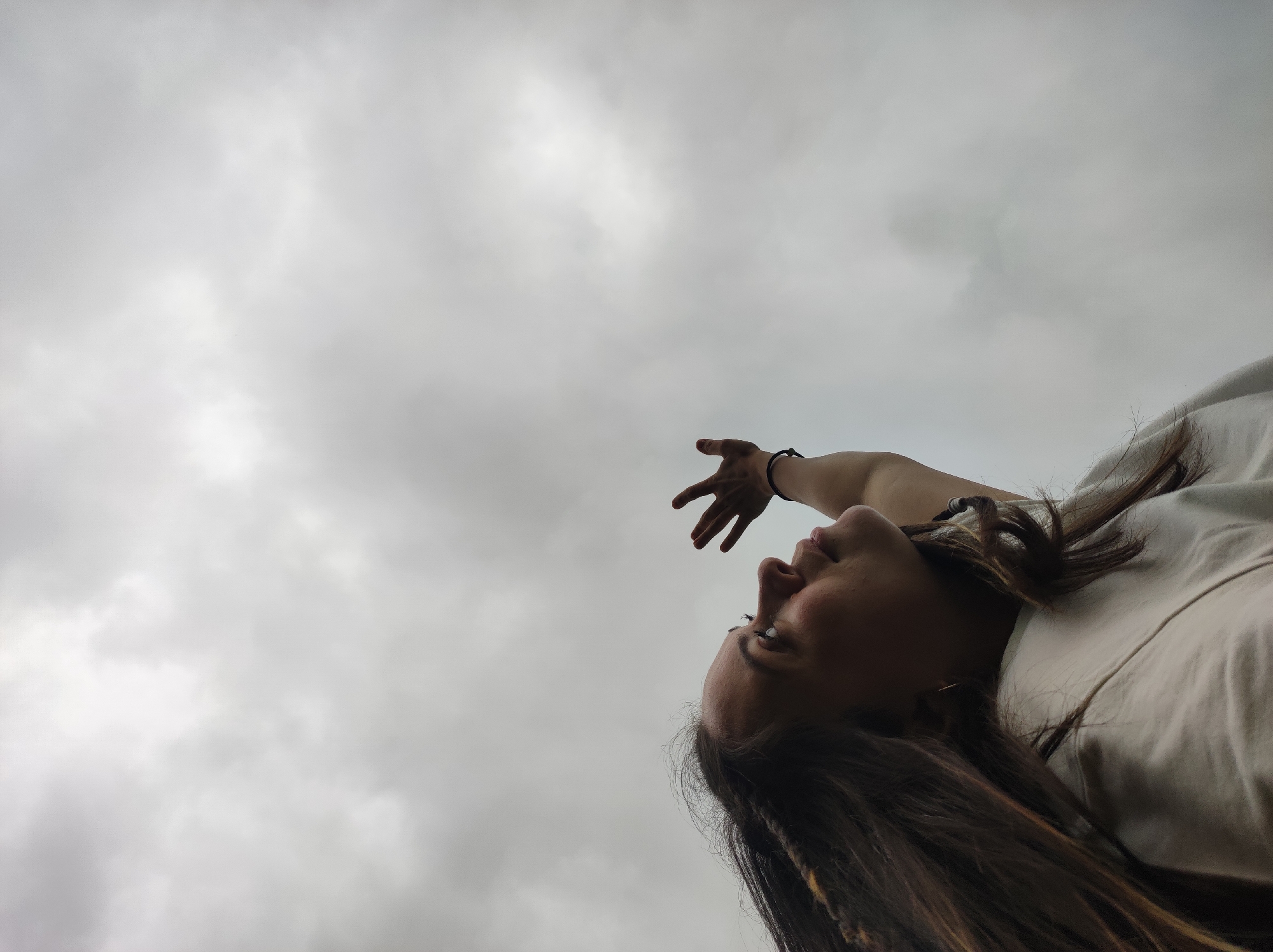 A low-angle shot captures a person, likely female, with long, dark hair, against an overcast sky in Фоки, Russia. The individual is positioned in the lower right portion of the frame, lying down or reclined, and gazing upwards. Their right arm is extended diagonally towards the upper left, with the hand open as if reaching. A dark band is visible on the wrist. The background is composed entirely of a cloudy, grey-white sky, indicating an overcast day.