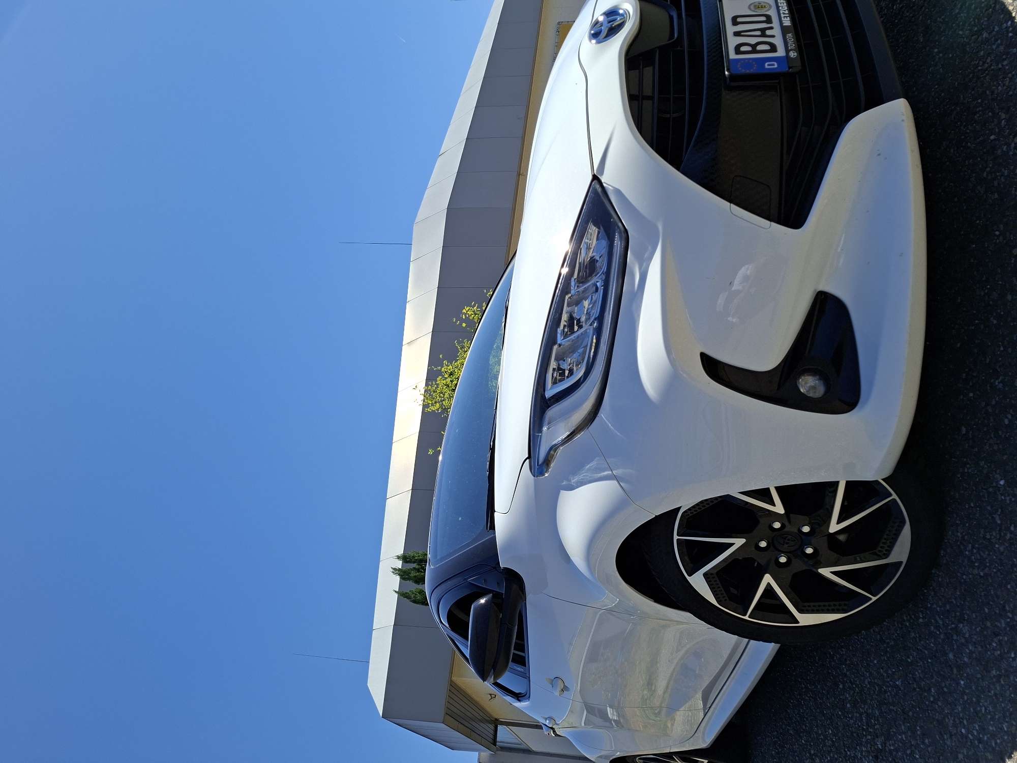 A white Toyota Yaris is parked outdoors on an asphalt surface, angled towards the viewer. The car is a hybrid model, as indicated by the blue accents on the Toyota emblem on the front grille. The license plate, visible on the front of the vehicle, reads "BAD 123" and includes the German country code "D" and the text "TOYOTA METZGER", suggesting the car is registered in or associated with Baden-Baden, Germany. The car's front right wheel is prominently displayed. In the background, a modern, light-colored building with a geometric design is visible, and the sky is clear and bright blue.