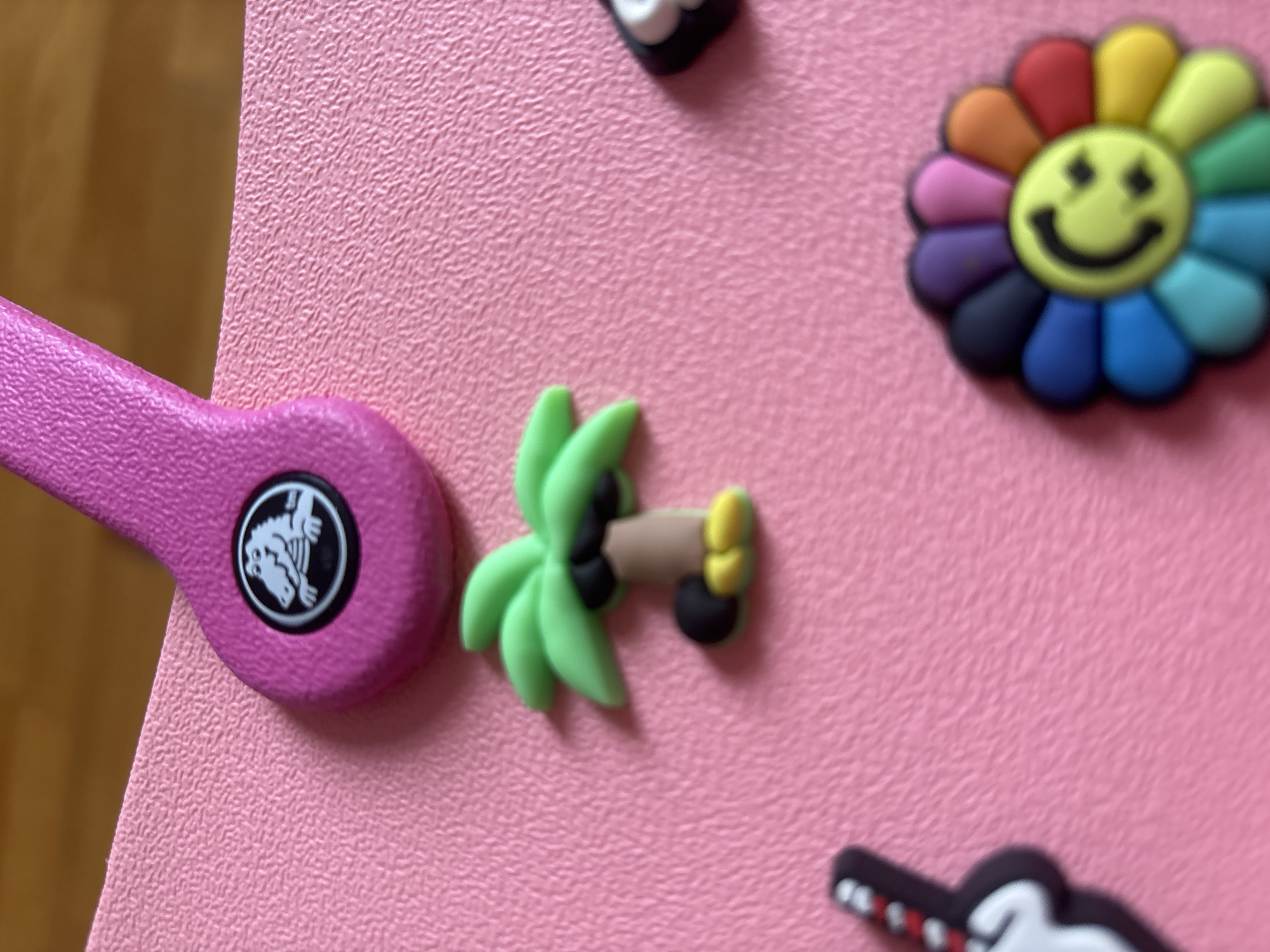 The image is a close-up, eye-level shot of a pink textured surface, likely a bag or accessory, adorned with several colorful charms. In the upper portion, a bright pink strap with a black circular emblem featuring a white alligator is visible. Below this, a small, green palm tree charm with a brown trunk and black and yellow accents is positioned slightly off-center to the left. To the right of the palm tree, a portion of a black and white charm, possibly a shoe or a face, is visible. Further down and to the right, a vibrant rainbow-colored flower charm with a yellow smiley face at its center is prominently displayed. In the lower left corner, a partially visible charm resembling a black and white guitar with red accents is present. The background is out of focus, showing a hint of a wooden surface. There are no people or animals in the image, and no specific location in Georgiyevsk, Russia can be determined from the provided visual information.