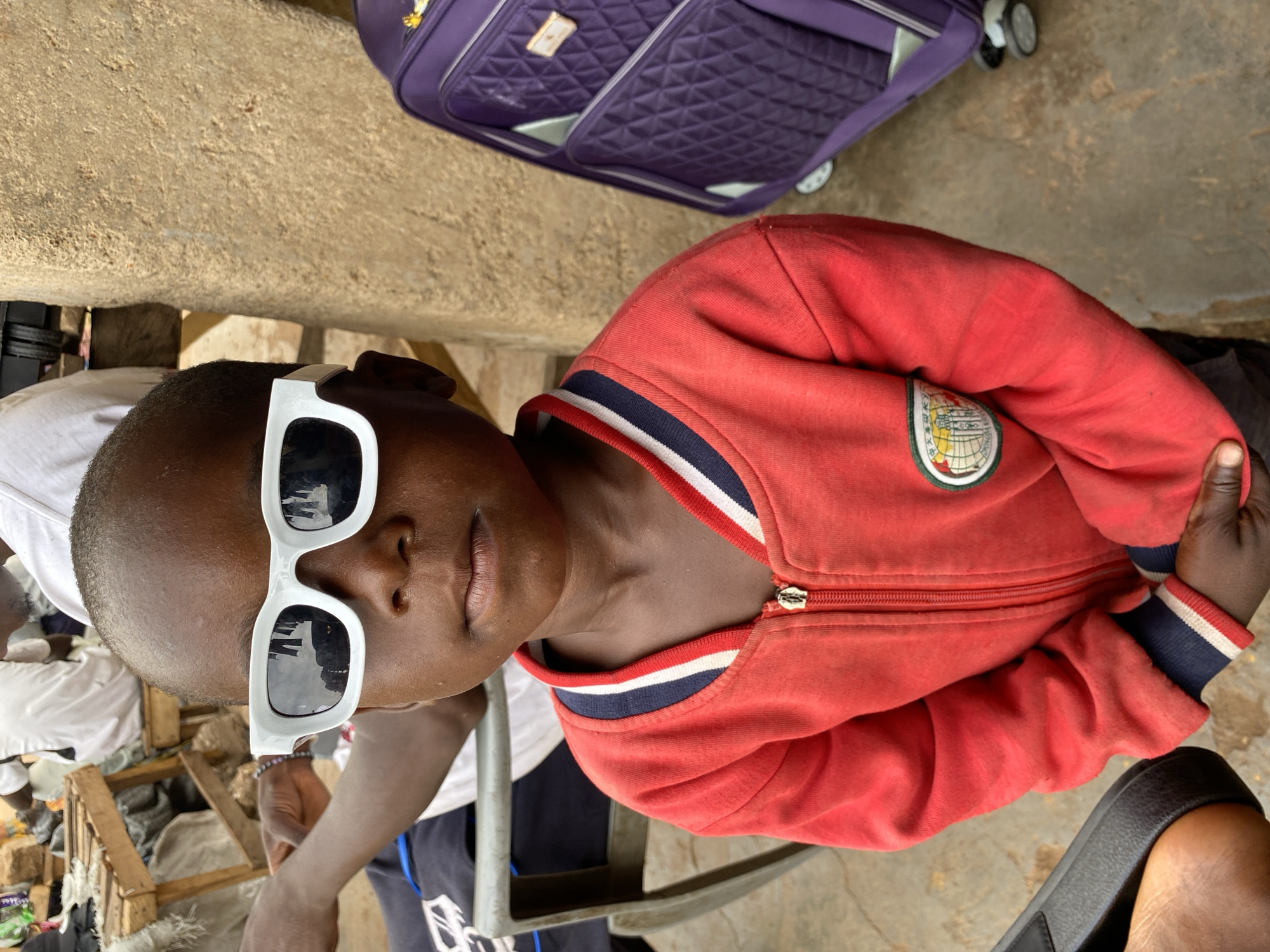 A child wears oversized white-framed sunglasses and a red zippered jacket with a school crest. The jacket has striped trim at the collar and cuffs. A purple quilted suitcase with wheels is visible in the background, suggesting travel. The child is positioned near an outdoor setting with concrete surfaces. The background includes a glimpse of what seems to be an informal outdoor marketplace or storage area. Bukuru, Nigeria, is not visually confirmed within this image.