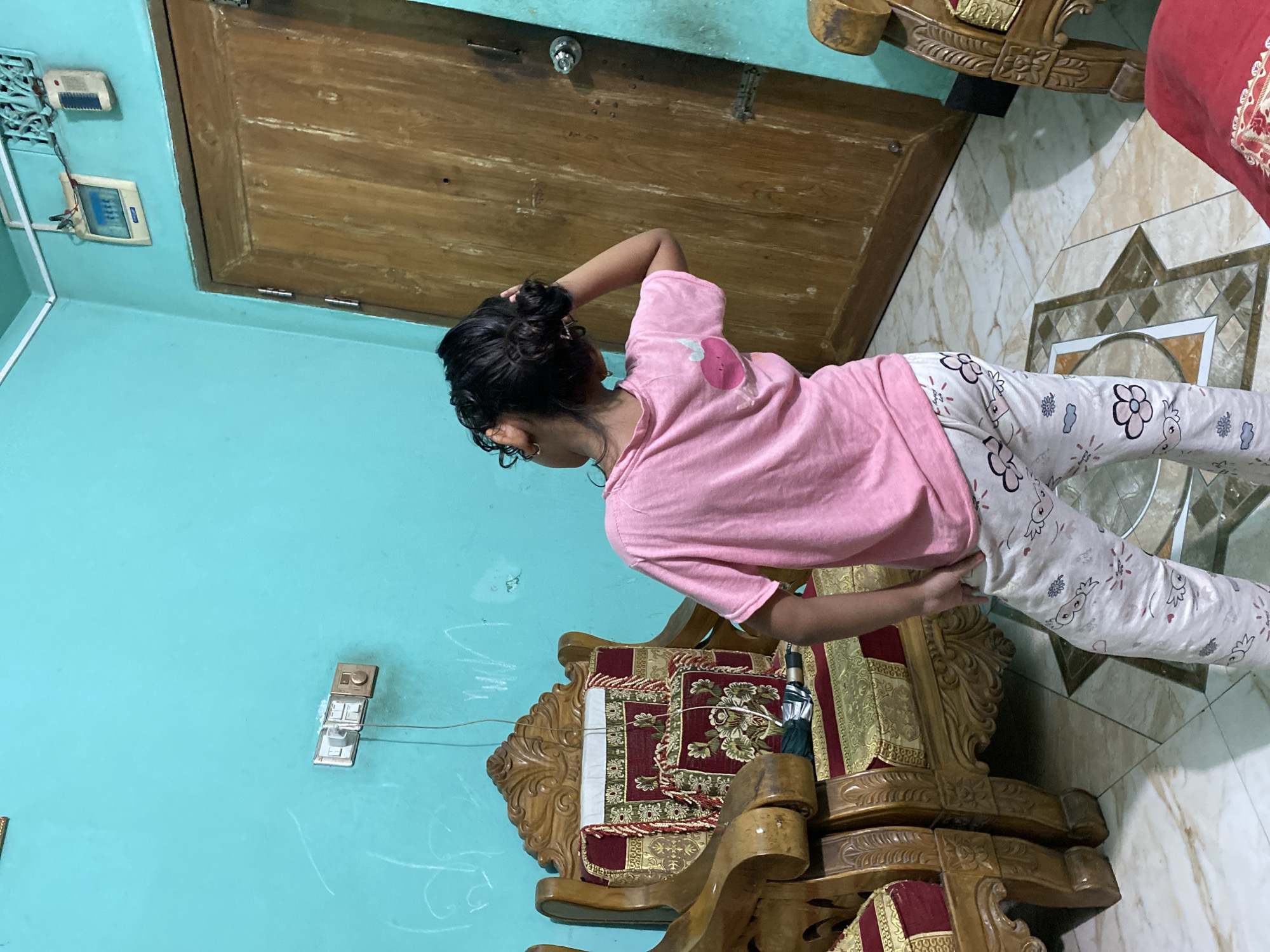 A young girl, with her back to the camera, stands in a room with teal-colored walls. She has dark, curly hair pulled up in a bun and is wearing a pink t-shirt and patterned pajama pants. The girl is looking up and to her right, with her right hand touching her head. To her left is a large, ornate wooden chair with red and gold patterned upholstery. To her right is a wooden door with a round doorknob. Electrical outlets and switches are visible on the wall. The floor is tiled with a geometric pattern. The overall impression is of a domestic interior in Dhaka, Bangladesh.