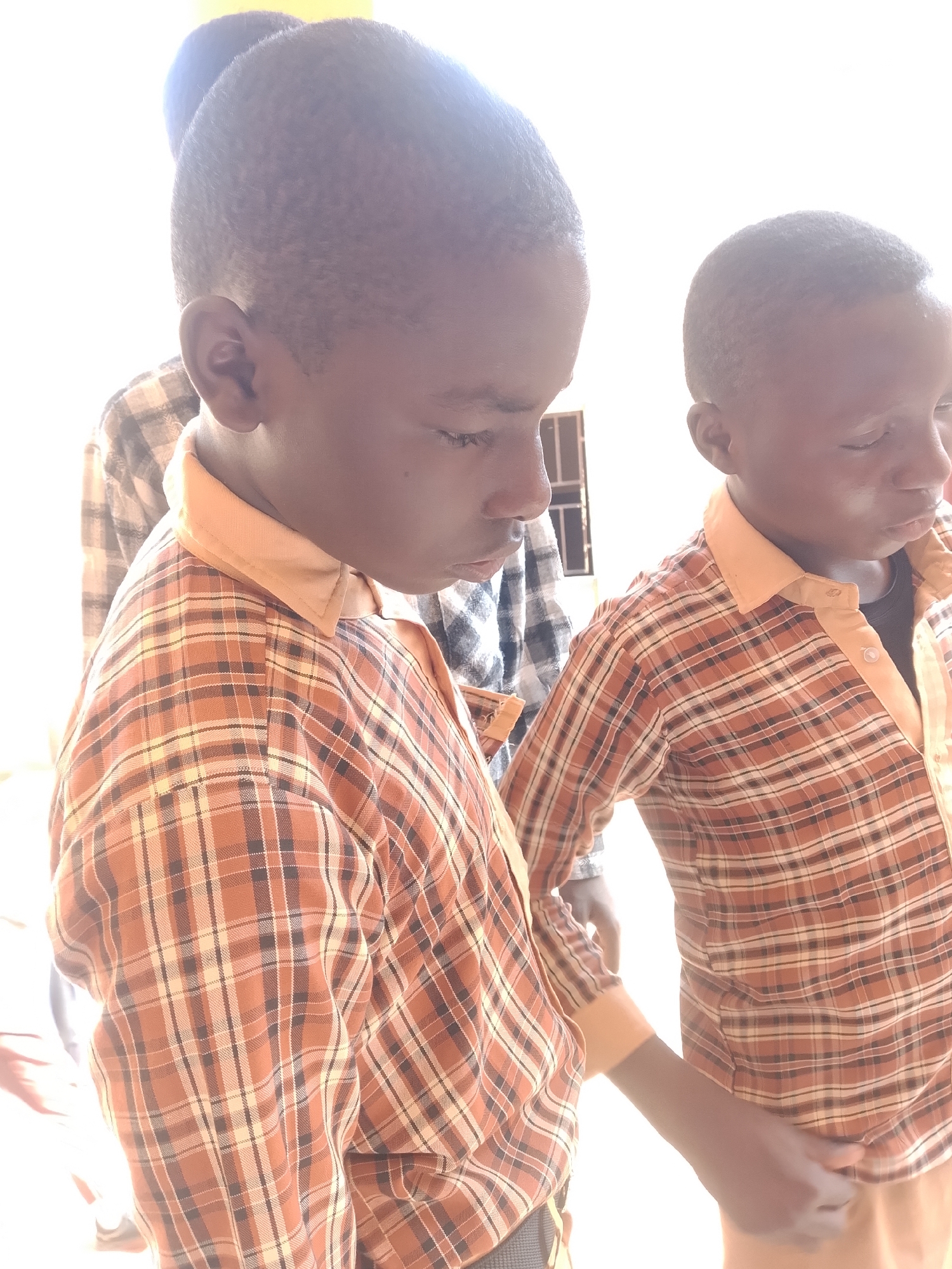 Two young Black boys, wearing plaid shirts, are standing close together. The boy in the foreground is looking down and to the left, his right hand is visible near his stomach. The boy to his right is also looking down and to the left, his right hand is near his chest. A third boy, mostly out of frame, is partially visible behind them to the left. They appear to be indoors, with a bright light source illuminating the scene from the right. The background is a plain white wall with a window frame visible on the right.