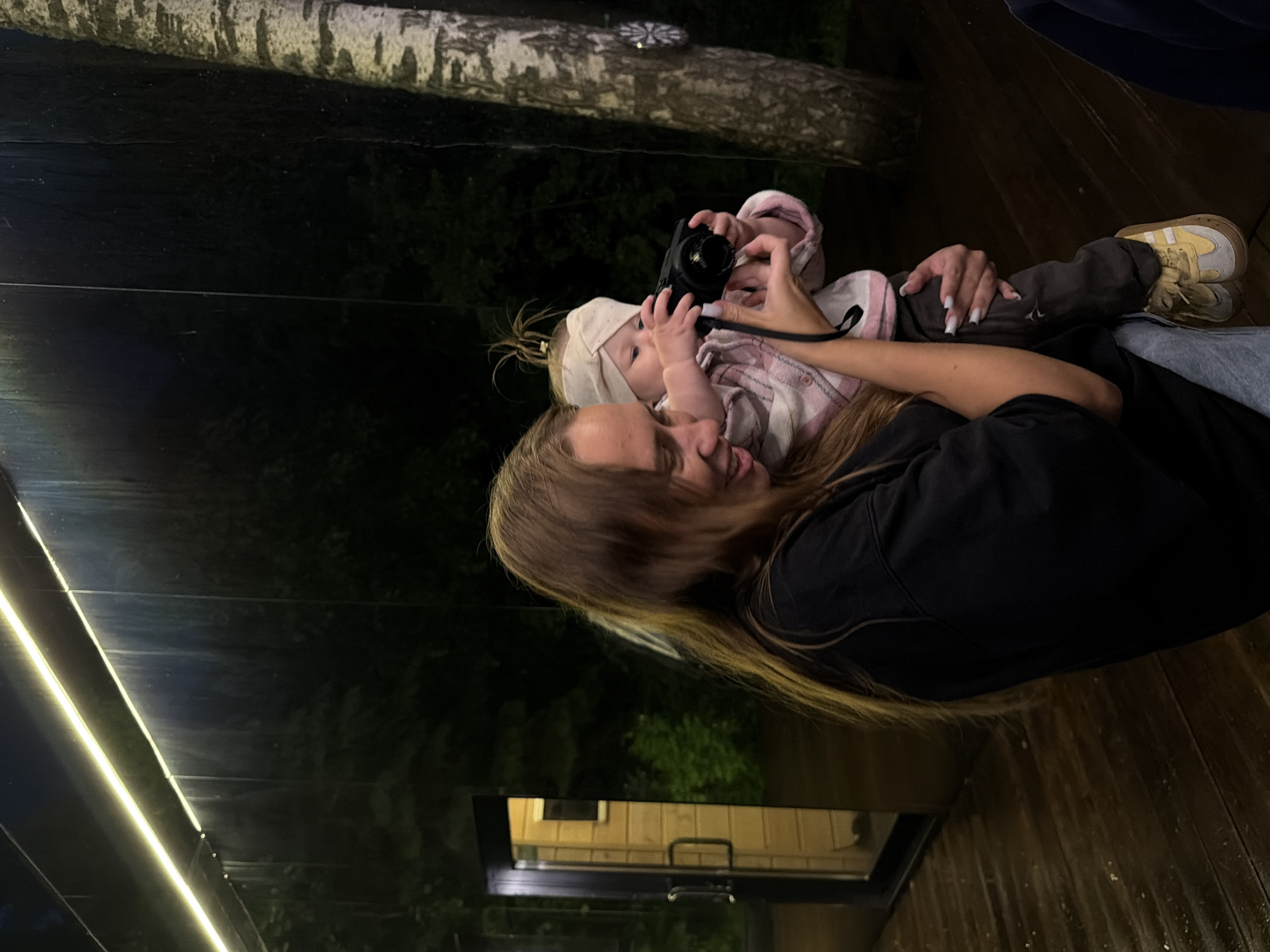 The image shows a woman holding a baby, who is playing with a compact camera. The woman is wearing a black top, and the baby is wearing a pink and white plaid shirt and a white head covering. The baby is holding the camera with both hands. The setting appears to be outdoors on a wooden deck, possibly in the city of Bakhtiyary, Russia, though this cannot be definitively confirmed from the image alone. The background suggests an outdoor environment with trees and possibly a building visible through a window. A string of lights is visible in the upper left of the frame.