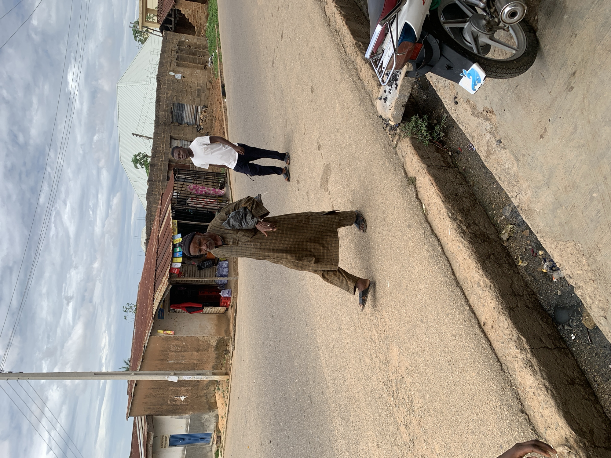 This image depicts an urban street scene in Jos, Nigeria, under an overcast sky. The foreground shows an asphalt road with a concrete drainage ditch running along its right edge, containing litter and dark liquid. The rear section of a white and red motorcycle, featuring a chrome rack and a dark mudguard with a partial blue and white sticker, is visible on the far right.
Two males are present on the road. In the left-midground, an older male with dark skin and a beard walks from left to right, dressed in a full-length, brown plaid garment and dark sandals, carrying a dark item in his left hand. In the midground-right, a younger male with dark skin walks towards the viewer, wearing a white t-shirt, dark trousers, and sandals.
Along the left side of the street, a tall concrete utility pole supports multiple overhead power lines. Behind it, a light-colored building with a blue door and a corrugated metal roof is visible, followed by an earth-toned building with a reddish corrugated metal roof, appearing to be a small shop with merchandise visible inside and colorful banners hanging from its eaves. On the right side of the street, beyond the drainage ditch, a partially constructed or deteriorated brick structure with a corrugated roof is present. Further in the background, a large building with a distinctive white, multi-sloped metallic roof is prominent against the cloudy sky.