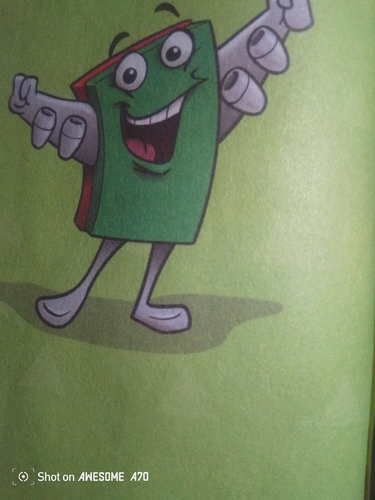 The image is a close-up shot of a cartoon character that resembles a green book with arms and legs, smiling broadly with its mouth open. The character has large, white eyes with black pupils and thin black eyebrows. It appears to be dancing or waving, with its arms extended and bent at the elbows, and its legs spread apart. The character is depicted against a plain, light green background. In the bottom left corner of the image, a watermark reads "Shot on AWESOME A70". There is no indication of the city of Jalingo, Nigeria, or any other specific urban location within the image. The image appears to be a photograph of a drawing or illustration.