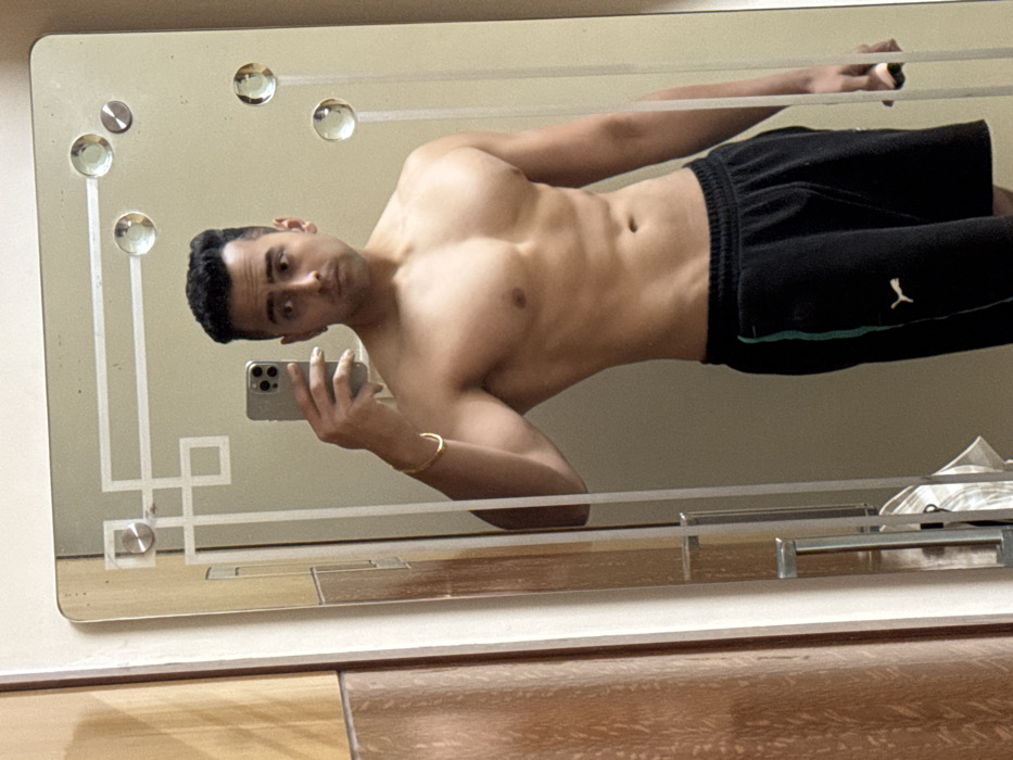 A male individual is captured in a mirror selfie within an indoor setting, likely a residential room. The subject, shirtless, displays a muscular physique and wears black athletic shorts featuring a green vertical stripe on the left side and a white Puma logo. He holds a silver-colored smartphone in his right hand, reflecting his face and the phone's camera lenses. A gold-colored bangle is present on his left wrist. The mirror, a prominent feature, is rectangular with etched geometric patterns and four circular glass embellishments. Reflected to the viewer's left, a wooden wardrobe or cabinet with two silver handles is partially visible. No specific visual elements within the image provide definitive information to determine a precise location within Kolkata, India.