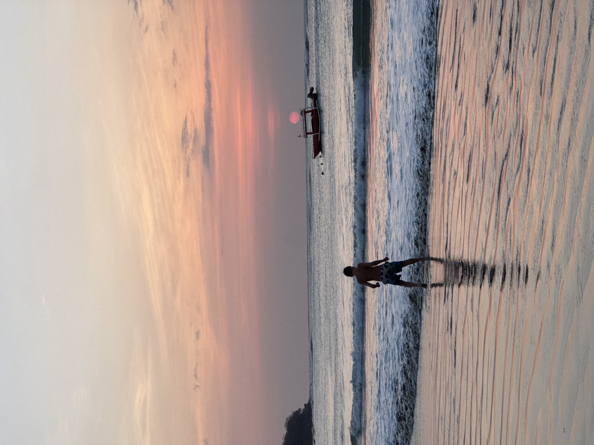 A young male, wearing patterned swim trunks, stands on a wet sandy beach, facing the ocean as a wave recedes. The beach is patterned with ripples from the receding water, reflecting the colors of the sunset. In the distance, a small red boat with an outboard motor is anchored offshore, with the sun setting behind it, casting a pinkish glow across the sky and the water. A headland covered in vegetation is visible in the far left of the image. The scene is set during sunset, likely on a beach in Kuta Selatan, Indonesia, given the commonality of such views and boat types in the region.