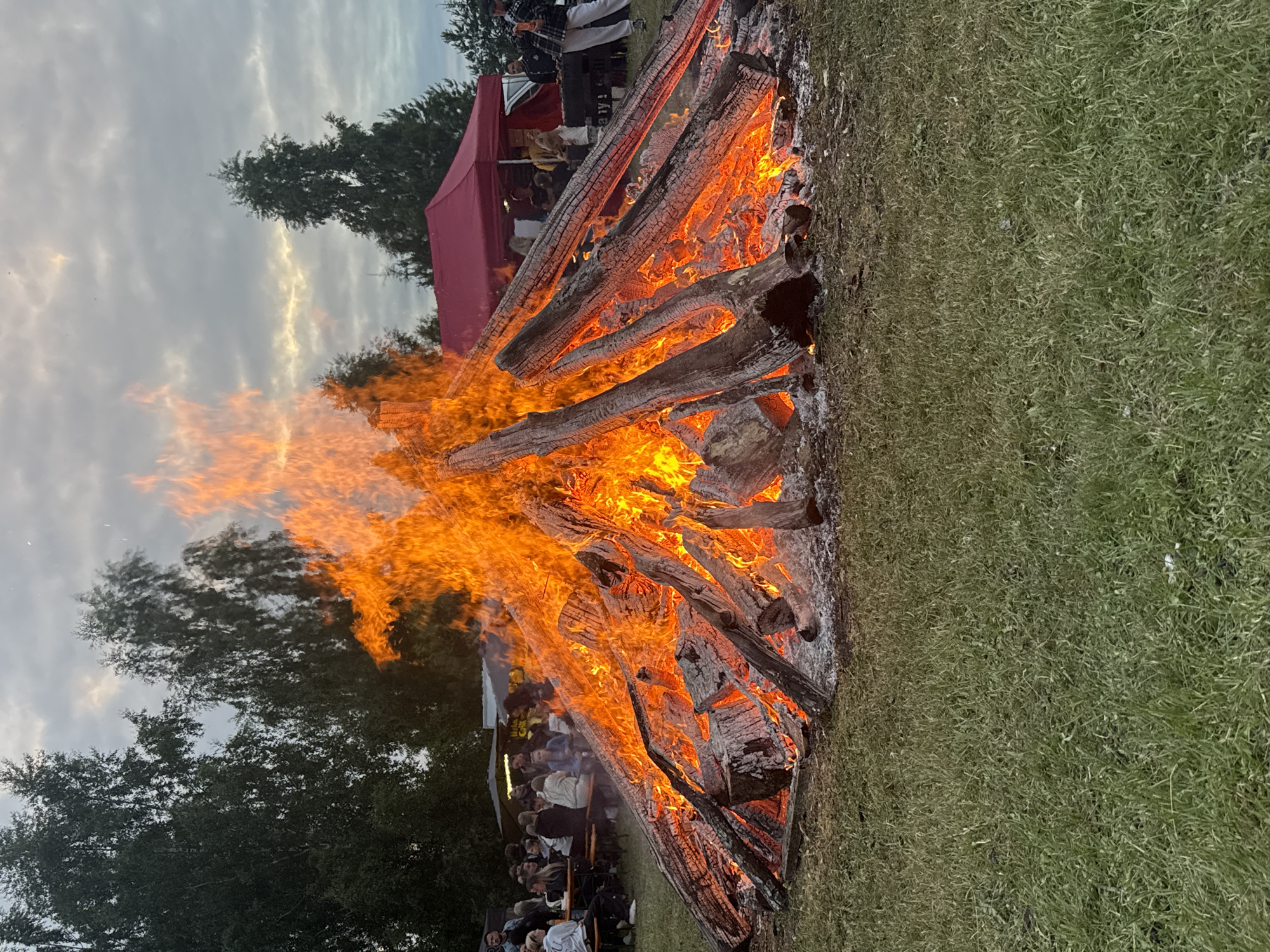 A large bonfire is burning on a grassy field, with logs of varying sizes fueling the flames. The fire is intense, with bright orange and yellow flames reaching upwards. In the background, a group of people are gathered, sitting on chairs and benches, watching the bonfire. There is also a red tent or canopy visible behind the crowd. Trees can be seen on the left side of the image. The sky is overcast with some patches of light, suggesting it is late afternoon or early evening. The overall mood appears to be one of communal gathering and enjoyment around the fire. This scene is taking place in Rapla, Estonia.
