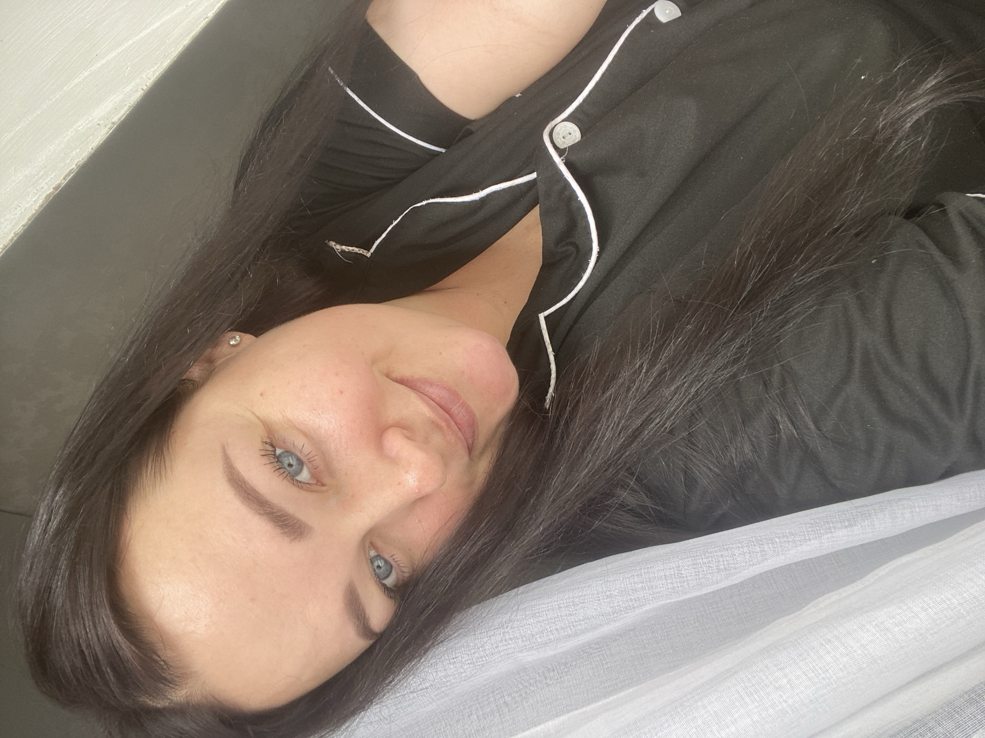 A woman with long, dark hair and striking blue eyes is the central subject of this close-up image. She is captured from the chest up, lying on her side and looking directly towards the camera with a soft, content smile. She is wearing black pajamas detailed with white piping and white buttons. A small, round, sparkly stud earring is visible in her left earlobe. The background consists of a plain, dark surface on the upper left and a soft, sheer white fabric, likely a curtain or part of the bedding, on the lower right. The lighting suggests an intimate indoor setting, contributing to the serene and relaxed atmosphere. Her expression conveys a sense of peacefulness and comfort, typical of a cozy moment at home in Sochi, Russia. No visible text is present in the image.