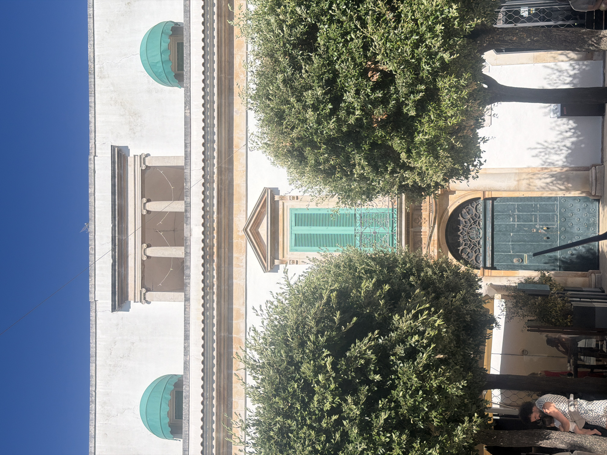 The image depicts a sunny street scene in Alberobello, Italy, focusing on the facade of a traditional building with a large, ornate green door and an arched entryway. Two lush green trees frame the entrance, their branches casting dappled shadows on the white wall behind them. On the left side, a man with curly hair is holding a baby. He is standing near the trunk of a tree. In the background, a woman is visible near a table, and another person is seated. Above the ground-floor entrance, a balcony with a turquoise shuttered window is visible, flanked by architectural elements that include columns and decorative awnings. The sky above is a clear, bright blue.