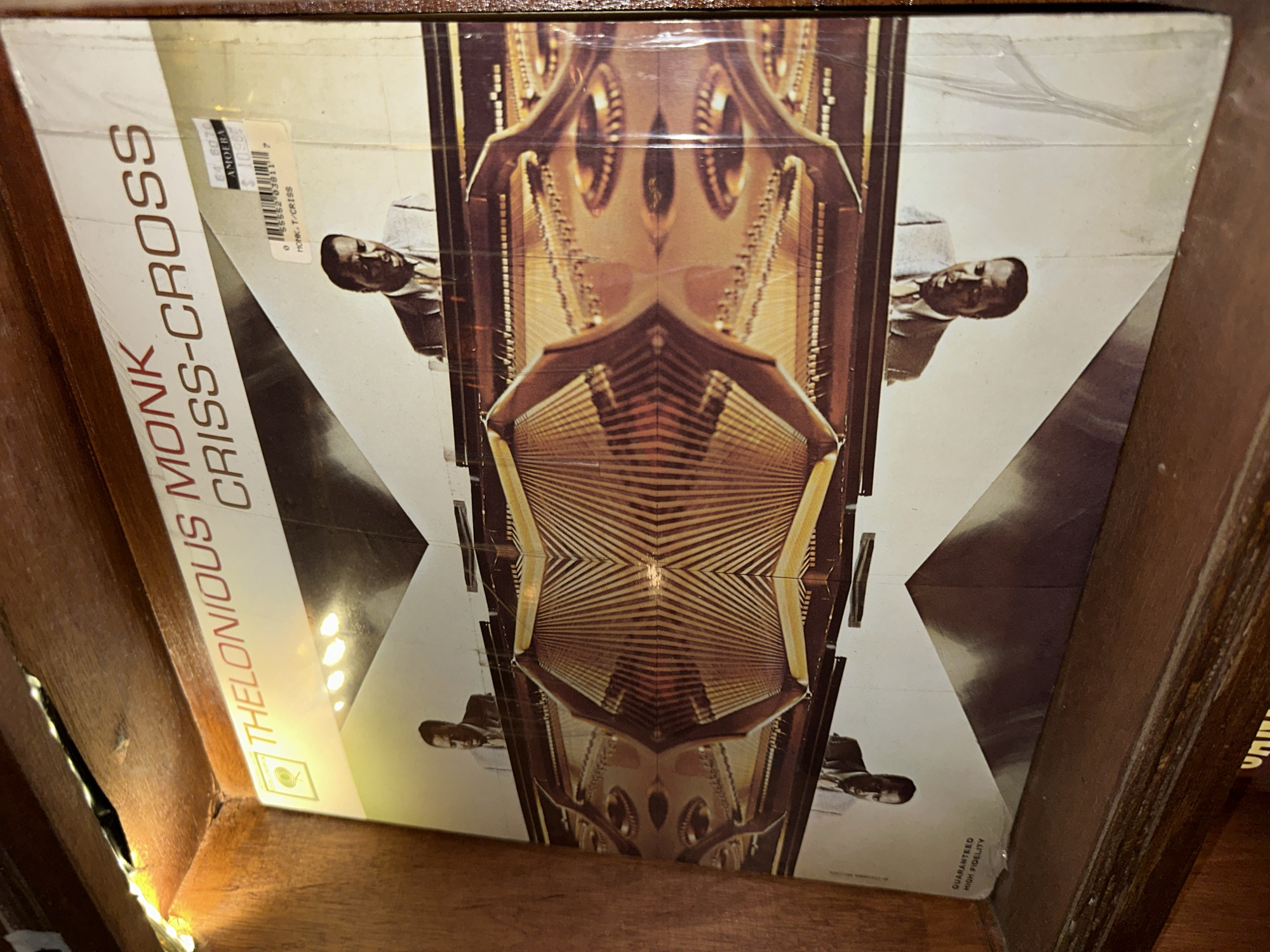 The image shows a vinyl record album titled "Thelonious Monk Criss-Cross" resting on a wooden shelf. The album cover features a stylized photograph of Thelonious Monk playing a piano, with the image mirrored to create a symmetrical effect. A price sticker is visible on the album cover. The album is part of a collection of records stored on wooden shelves, illuminated by fairy lights along the top edge of the shelf. The environment appears to be indoors, possibly a record store or a personal collection. The specific location in Davao City, Philippines, cannot be determined from the visual information provided in the image.