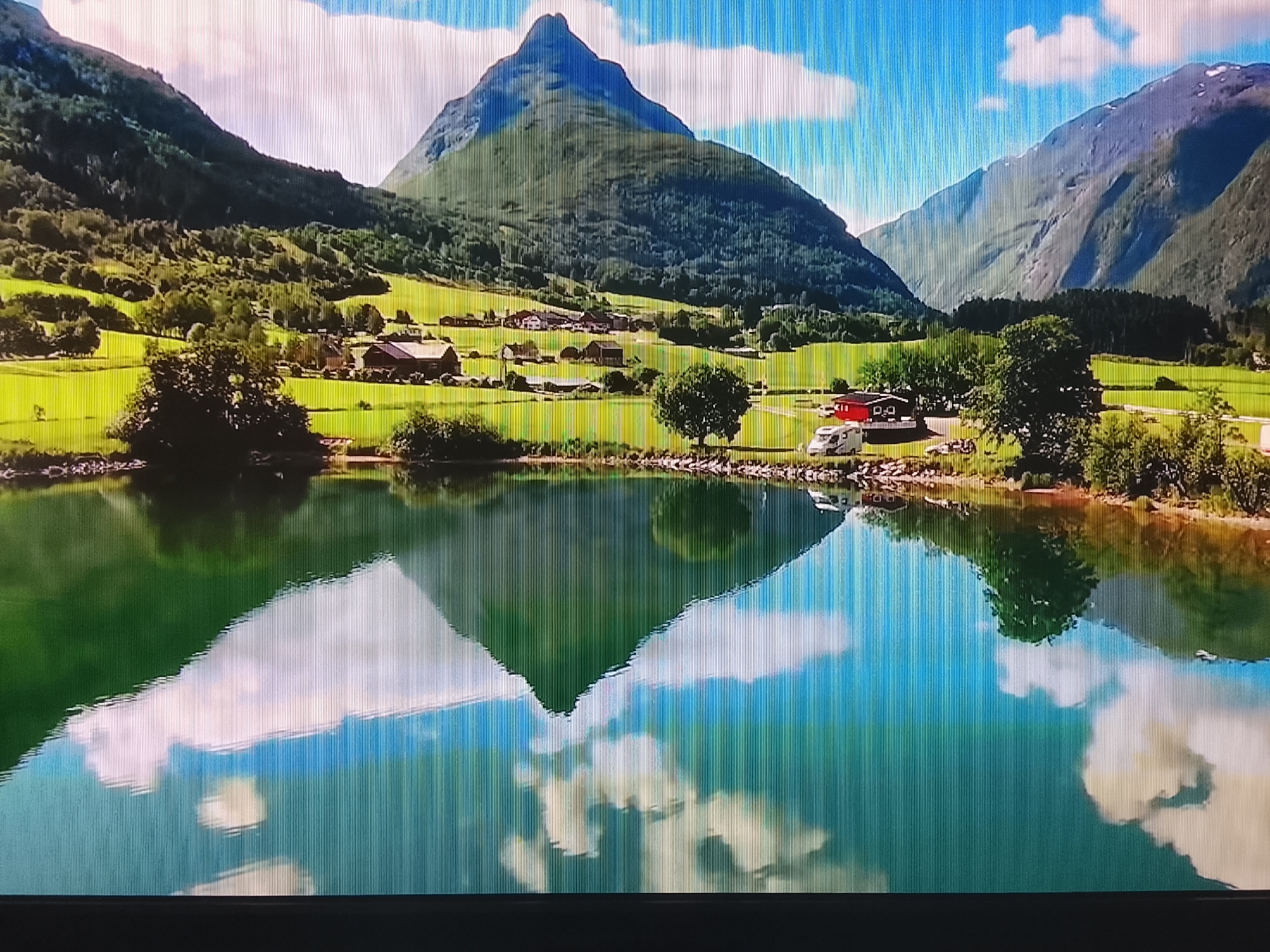 This is a wide-angle, landscape photograph of a serene body of water reflecting a mountainous region. The scene is set in a rural environment with a lake in the foreground, and lush green hills and a prominent, conical mountain in the background. Several buildings, appearing to be houses or farm structures, are visible nestled on the slopes of the hills. A few trees dot the landscape. The sky is partly cloudy with a bright blue hue. The overall impression is one of natural beauty and tranquility. The image shows no people or any indications of urban life, and therefore, cannot be associated with the city of Ivanovo, Russia.