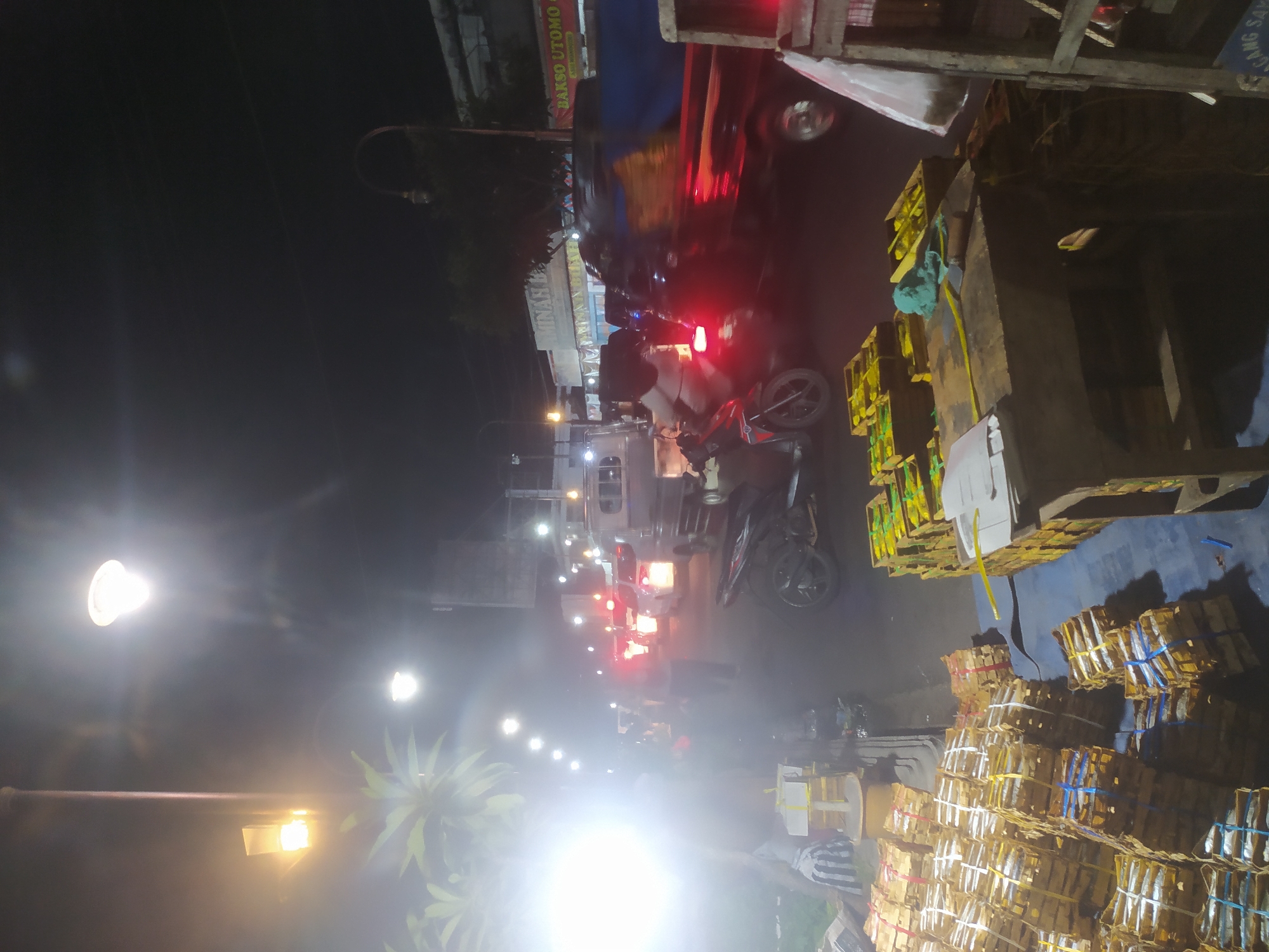 This is a nighttime scene captured on a city street in Kupang, Indonesia, likely a market or roadside stall area. The foreground is dominated by stacks of neatly arranged goods, appearing to be packaged items like crackers or snacks, bundled with colored string. To the right, a wooden stall structure is visible.
The street itself is illuminated by streetlights and the headlights and taillights of vehicles. A red motorcycle is parked in the middle ground, and a man is riding another motorcycle further down the street, partially obscured by motion blur. Several cars and a truck are also present, caught in traffic or moving along the road. The background shows buildings and more streetlights, with a general impression of a bustling urban environment at night. The overall lighting is dim and atmospheric, with bright light sources creating lens flares.