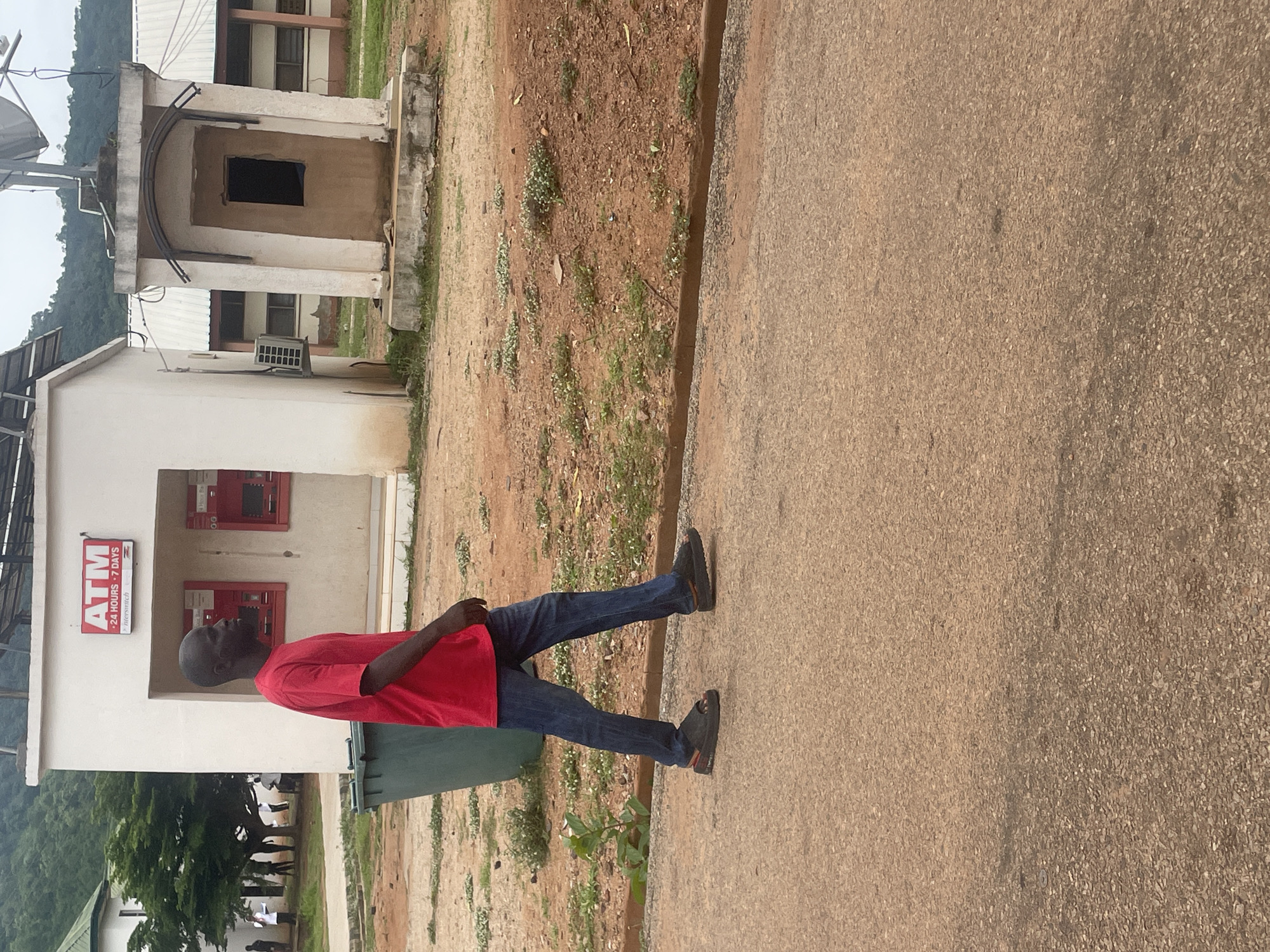A full shot shows a Black man in a red t-shirt and blue jeans walking from left to right in front of an ATM booth and a guard booth. The man's face is in profile, and he is wearing black sandals. The ATM booth is white with a red sign that reads "ATM" in white lettering and "24 HOURS 7 DAYS" in smaller white lettering. There are two ATM machines visible inside the booth. The guard booth is also white and has a small, dark window. In the background, there are trees, buildings, and a hill. The ground is a mix of pavement and dirt. A green trash can is visible to the left of the ATM booth.
