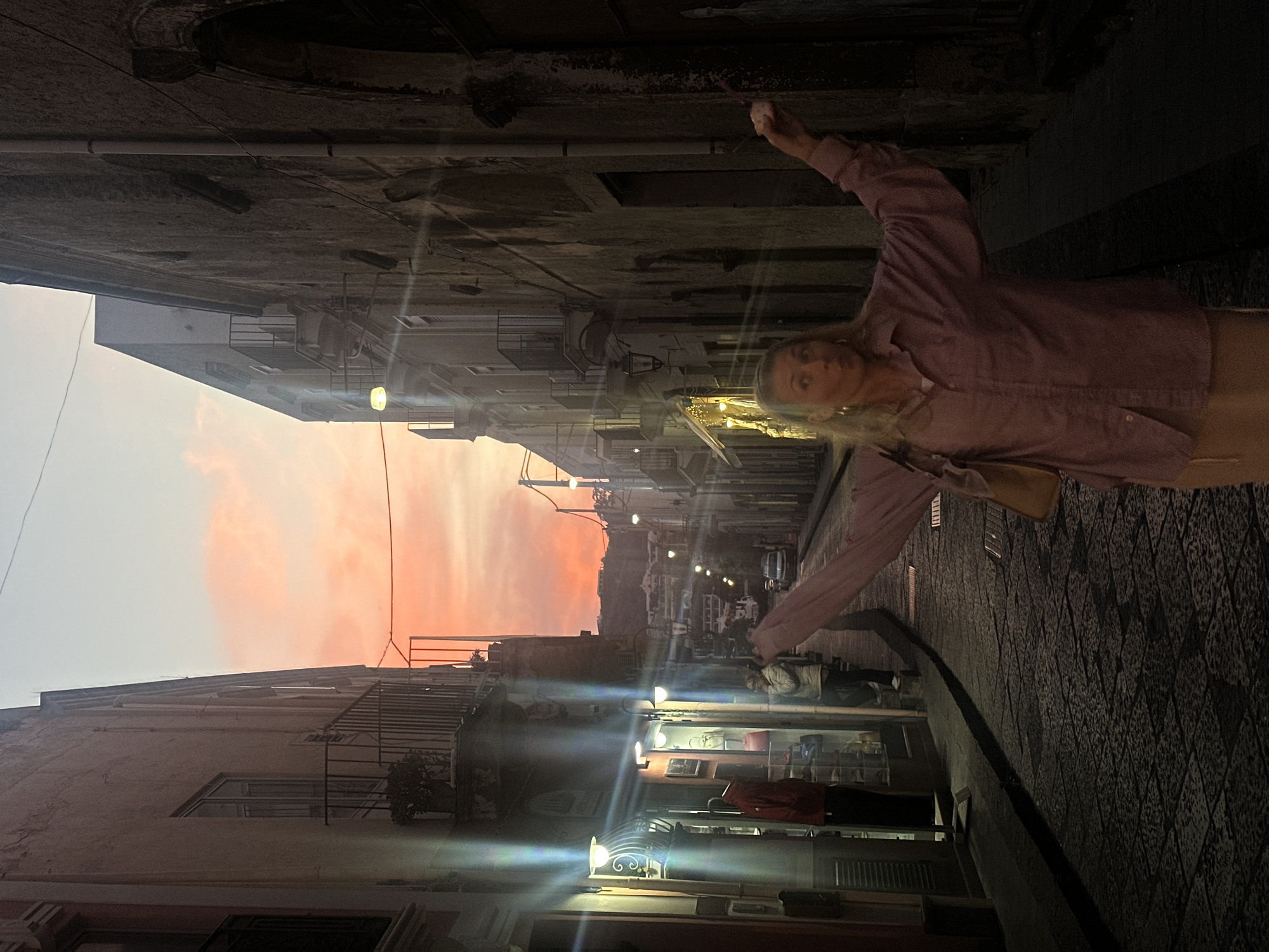 The image depicts a narrow street in Lipari, Italy, at dusk or early evening, seen from a perspective looking up the street.
In the foreground, a blonde woman, wearing a light pink button-up shirt and light-colored pants, strikes a dynamic pose with her arms outstretched and raised, looking directly at the camera. A light-colored shoulder bag hangs from her left side.
The street is lined with multi-story, aged buildings on both sides. On the right side, several shopfronts emit bright artificial light, creating strong lens flares that streak across the image. Other individuals are visible further down the street, some appearing to walk. The street itself is paved with a distinctive patterned cobblestone or tiled surface. The undersides of balconies or upper stories overhang the narrow passage, revealing exposed pipes and wires.
Above the buildings in the distance, a vibrant sunset illuminates the sky with prominent hues of orange and pink, indicating the time of day is either dusk or early evening. The overall atmosphere is enhanced by the interplay of natural sunset colors and the artificial street and shop lighting. No discernible text is visible on any signs or buildings.