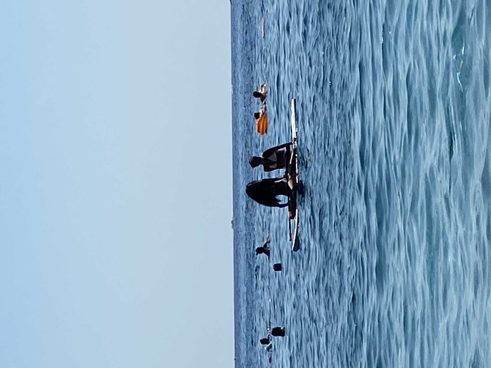 In the clear blue waters off the coast of Prijevor, Montenegro, several individuals are enjoying a day of recreation. Two people are seated on a paddleboard in the foreground. One individual with long dark hair is lying down on the paddleboard while the other person, wearing a dark top and a white bikini bottom, sits upright. Further out in the water, other people are swimming and floating. One person is on an orange inflatable float, and another is partially submerged. The horizon line is clear, with a faint hint of land or structures in the distance. The scene is bathed in bright sunlight, indicating a clear and warm day.