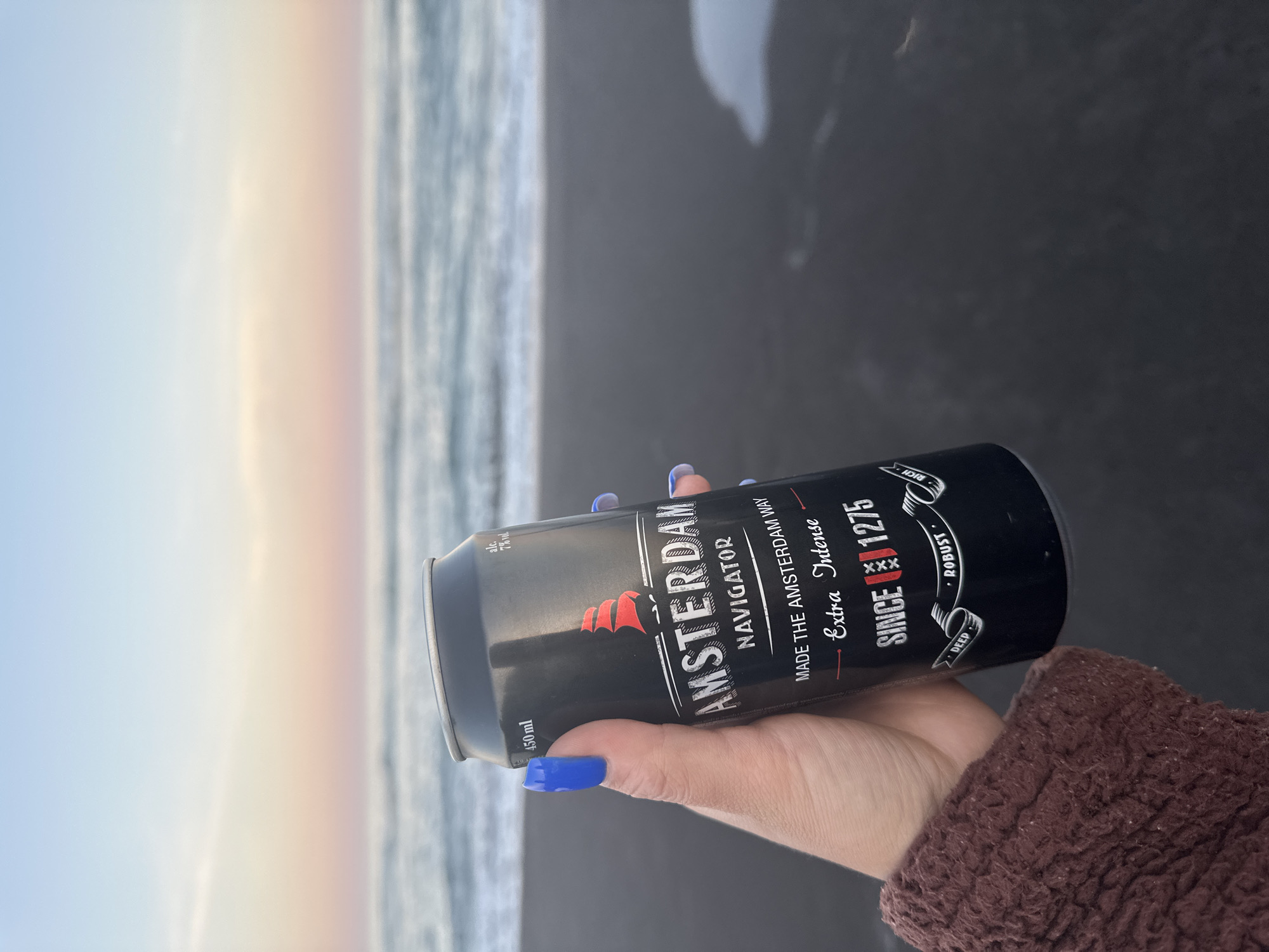 A person's hand with bright blue fingernails is holding a black can of Amsterdam Navigator beer on a dark sandy beach. The can has red lettering and a red ship logo. In the background, the ocean waves are gently rolling towards the shore under a pale pink and blue sky. The location is identified as Petropavlovsk-Kamchatsky, Russia.