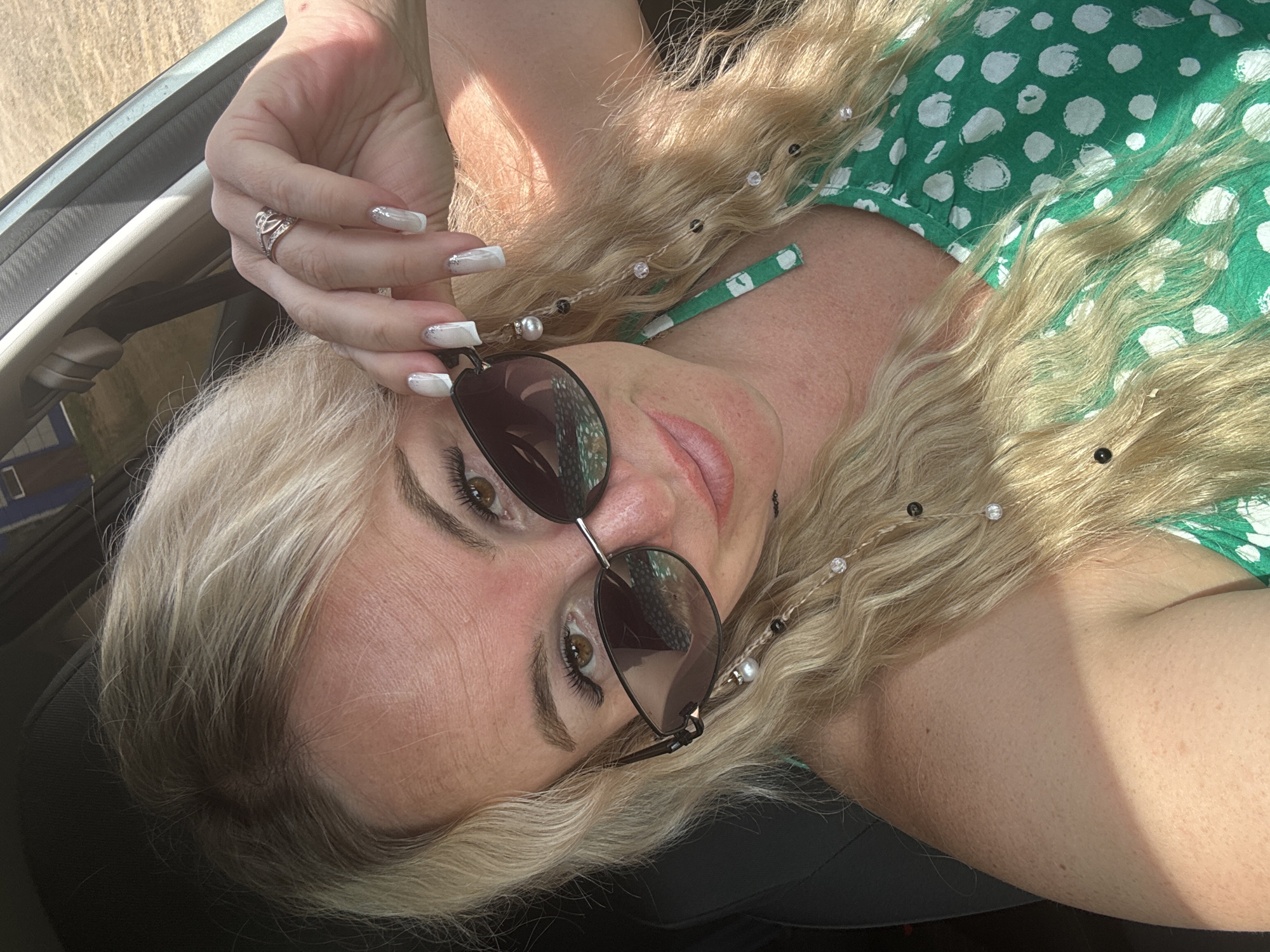 A blonde woman is pictured inside a car, possibly in Nozhovka, Russia, during the day. She is wearing sunglasses and a green and white polka dot dress. Her right hand is raised, and she is touching her sunglasses with her fingers. Her nails are painted white with silver glitter accents. She is also wearing rings on her fingers. The environment outside the car appears to be a rural or outdoor setting, with sunlight illuminating the scene. The car's interior is visible, with a dark-colored seat and part of the dashboard. Her hair is styled in loose waves, and her eyebrows are defined. The overall impression is a candid shot, possibly a selfie, taken in bright daylight.