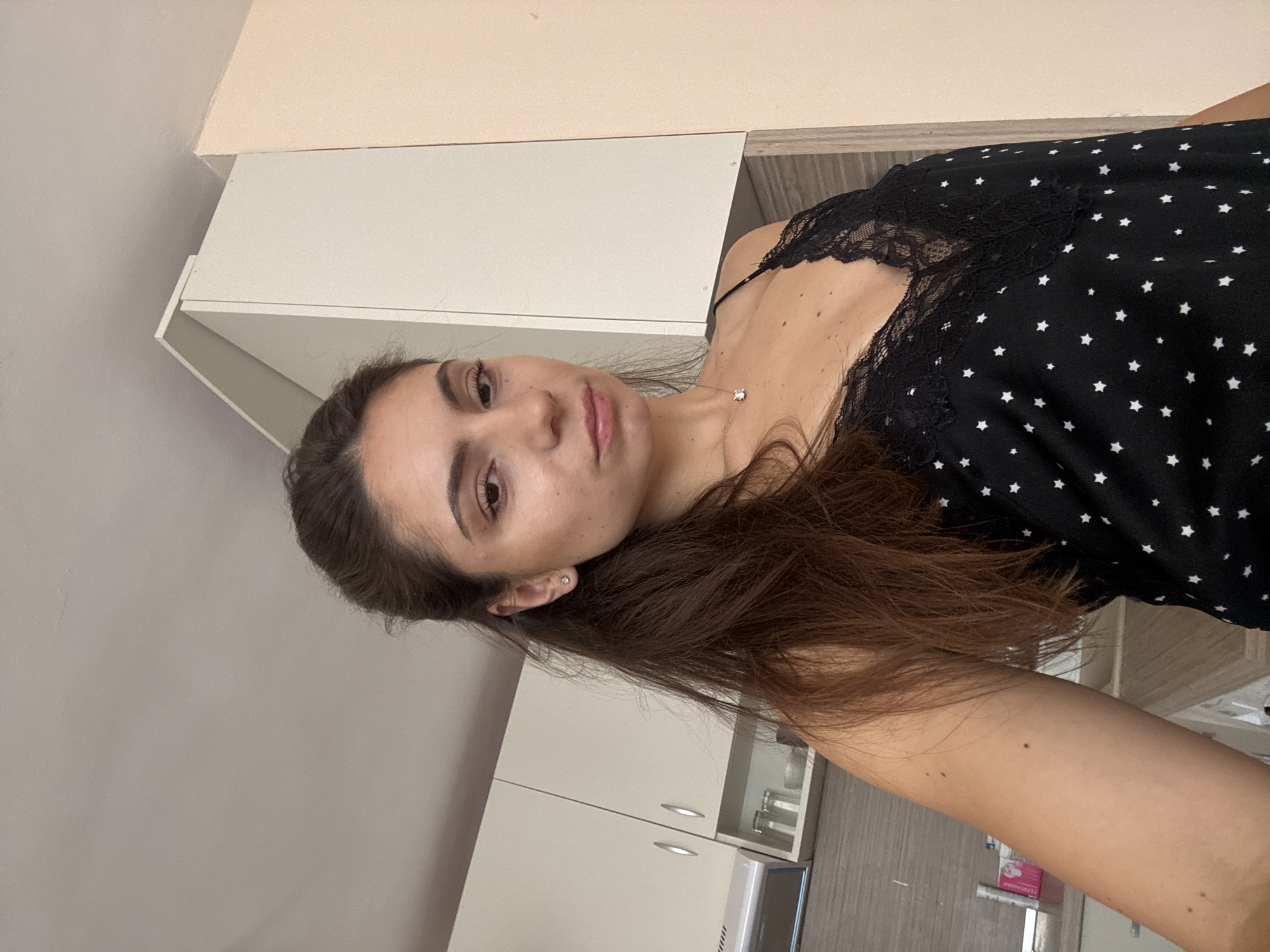 The image shows a medium shot of a young woman with long, dark brown hair, wearing a black camisole with white stars and a delicate necklace. She is standing in what appears to be a kitchen, with light-colored cabinets and countertops visible in the background. The scene is likely indoors, and the lighting suggests it could be daytime.