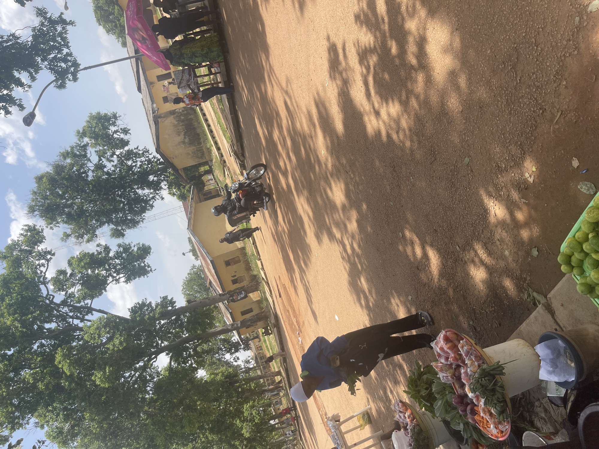 The image captures an outdoor market scene in Rafin Par, Nigeria. In the foreground, a person wearing a white headscarf and a blue jacket is bent over a collection of produce. To the left of this person, a white table displays some of the goods, including red onions, carrots, and leafy green vegetables. Further to the left, a large, shallow bowl overflows with fruits, possibly tomatoes and other round produce, all neatly arranged.
In the middle ground, a man wearing a helmet and a dark jacket is riding a motorcycle with a large cargo box attached to the back. The motorcycle is moving from left to right across the dusty, reddish-brown ground. Behind the motorcycle, a few other people are visible, some walking and others standing near stalls.
The background features a row of single-story buildings with yellow walls and corrugated metal roofs. Large, mature trees with dense green foliage line the street, casting dappled shadows on the ground. The sky is bright blue with scattered white clouds. An umbrella, possibly part of a market stall, is visible to the right of the frame, partially obscuring the buildings. The overall environment suggests a bustling day at a local market.