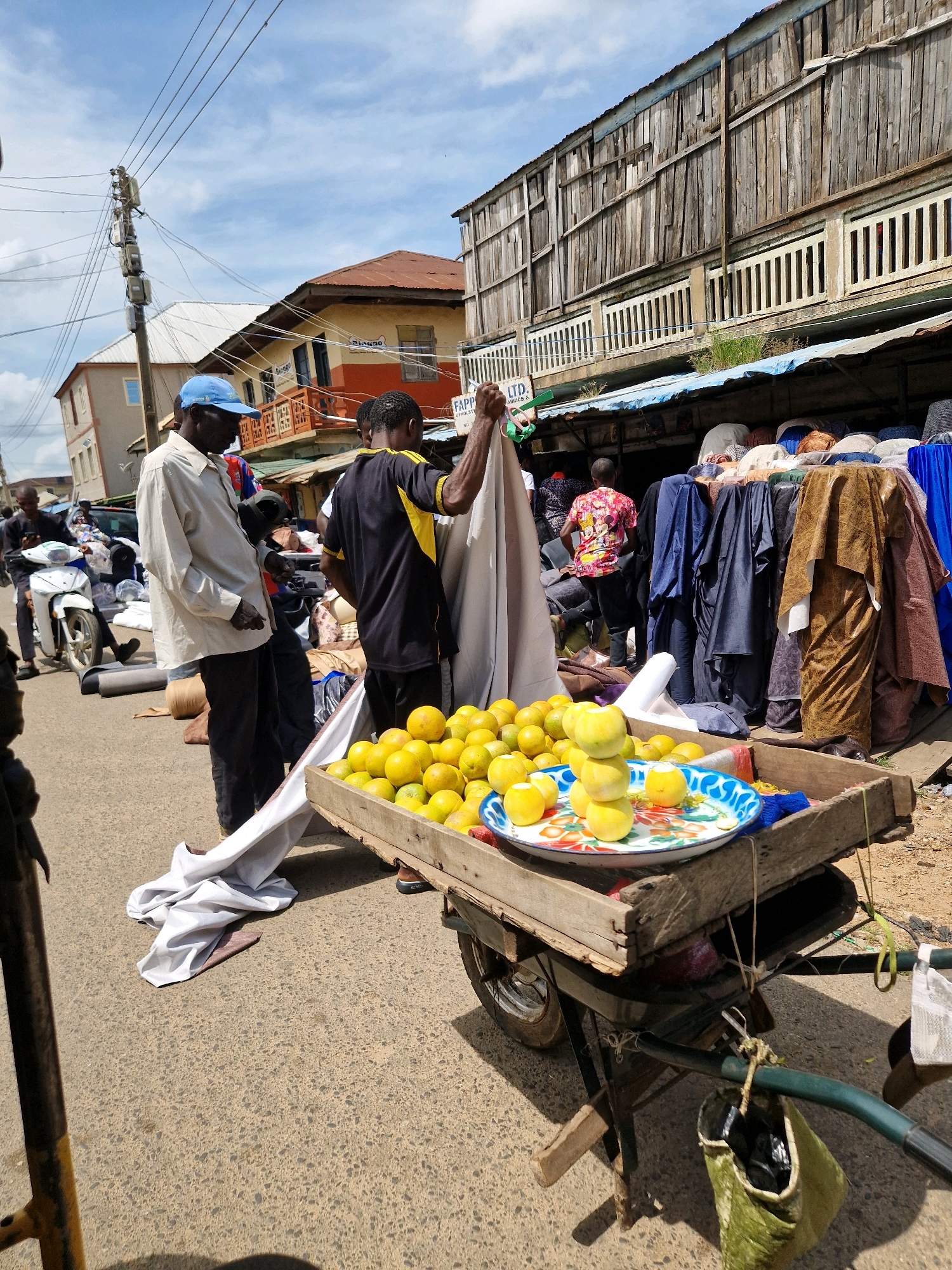 An outdoor market scene is depicted in Jos, Nigeria, under a partly cloudy sky. The foreground features a two-wheeled cart laden with produce, predominantly a pile of yellow-orange citrus fruits on a patterned tray. The cart's wooden construction is visible, along with a green bag attached to the handle. A man in a light-colored shirt and dark pants, wearing a blue cap, stands to the left, looking down. To his right, another man in a black and yellow shirt is seen from the back, holding a white cloth. Behind them, a bustling marketplace unfolds with stalls displaying various fabrics and clothing. Buildings with weathered wooden facades and corrugated iron roofs line the street. Several people are visible in the background, and a white motorbike is parked further down the road. The environment suggests a lively commercial area.