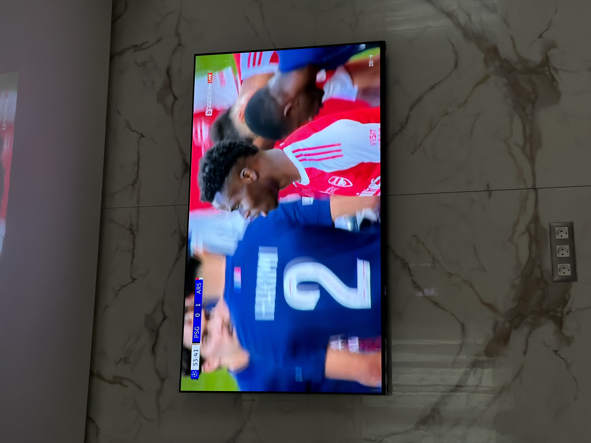A television displaying a soccer game is mounted on a marble-patterned wall. The soccer game's scoreboard indicates PSG 0, ARS 1 at the 53:41 mark. The soccer players are wearing red and blue uniforms. A European-style power outlet is visible on the wall to the right of the television. The location is likely indoors, but the image provides no additional information to place the image in Asenovgrad, Bulgaria.