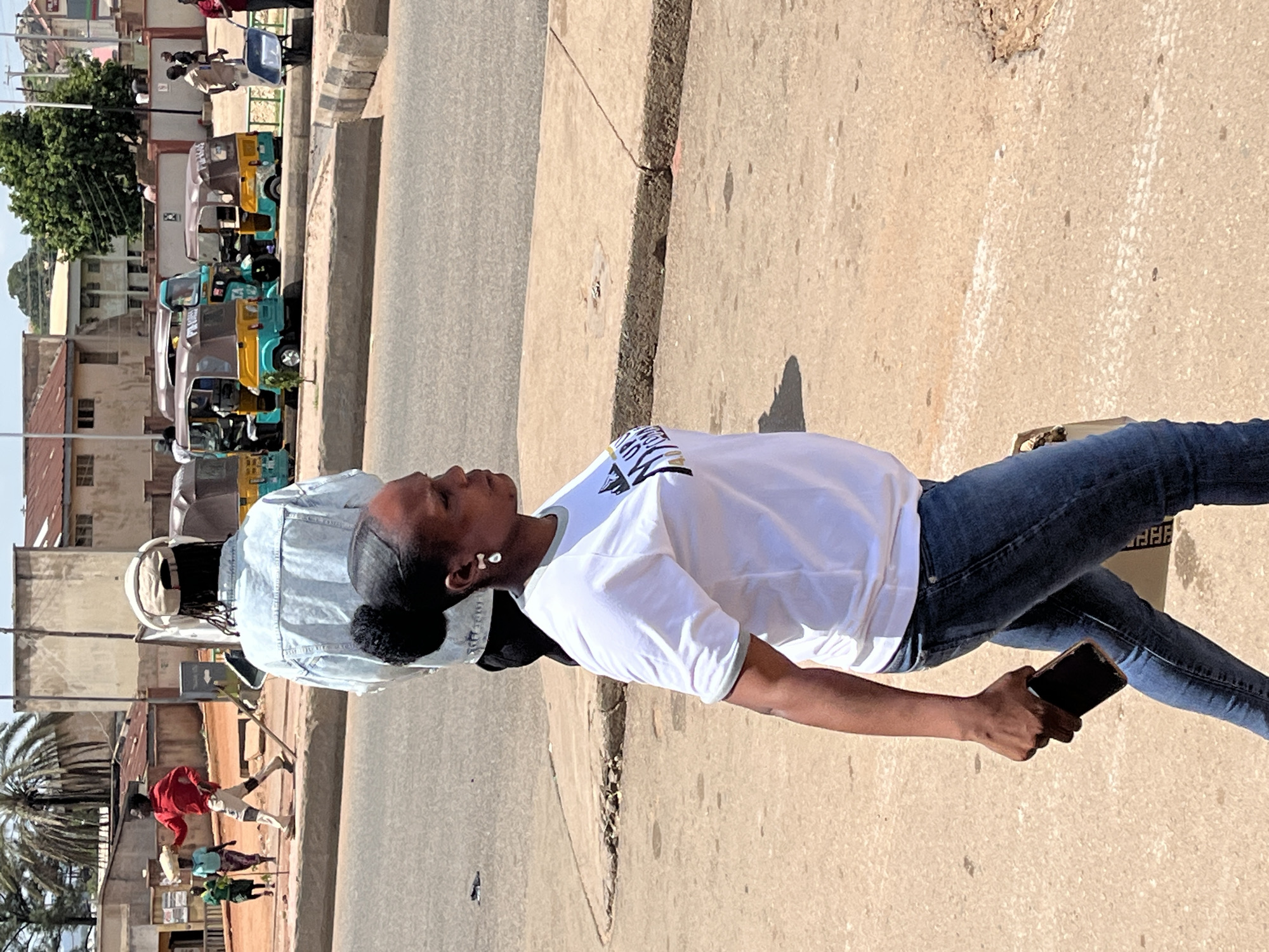 The image shows a street scene in Gura Top, Nigeria. In the foreground, a Black woman with her hair in a bun is walking to the right, looking away from the camera. She is wearing a white t-shirt with a graphic design and blue jeans. She is holding a dark-colored smartphone in her right hand. Behind her, a person wearing a light blue denim jacket and a beige cap is walking away from the camera. Further in the background, several auto-rickshaws, commonly known as "Keke NAPEPs," are parked or driving on the street. There are buildings and vegetation visible on either side of the street, suggesting an urban environment. Other people are also visible in the distance, including individuals carrying items and one pushing a wheelbarrow. The ground is paved with asphalt and concrete. The lighting suggests it is daytime with a clear sky.