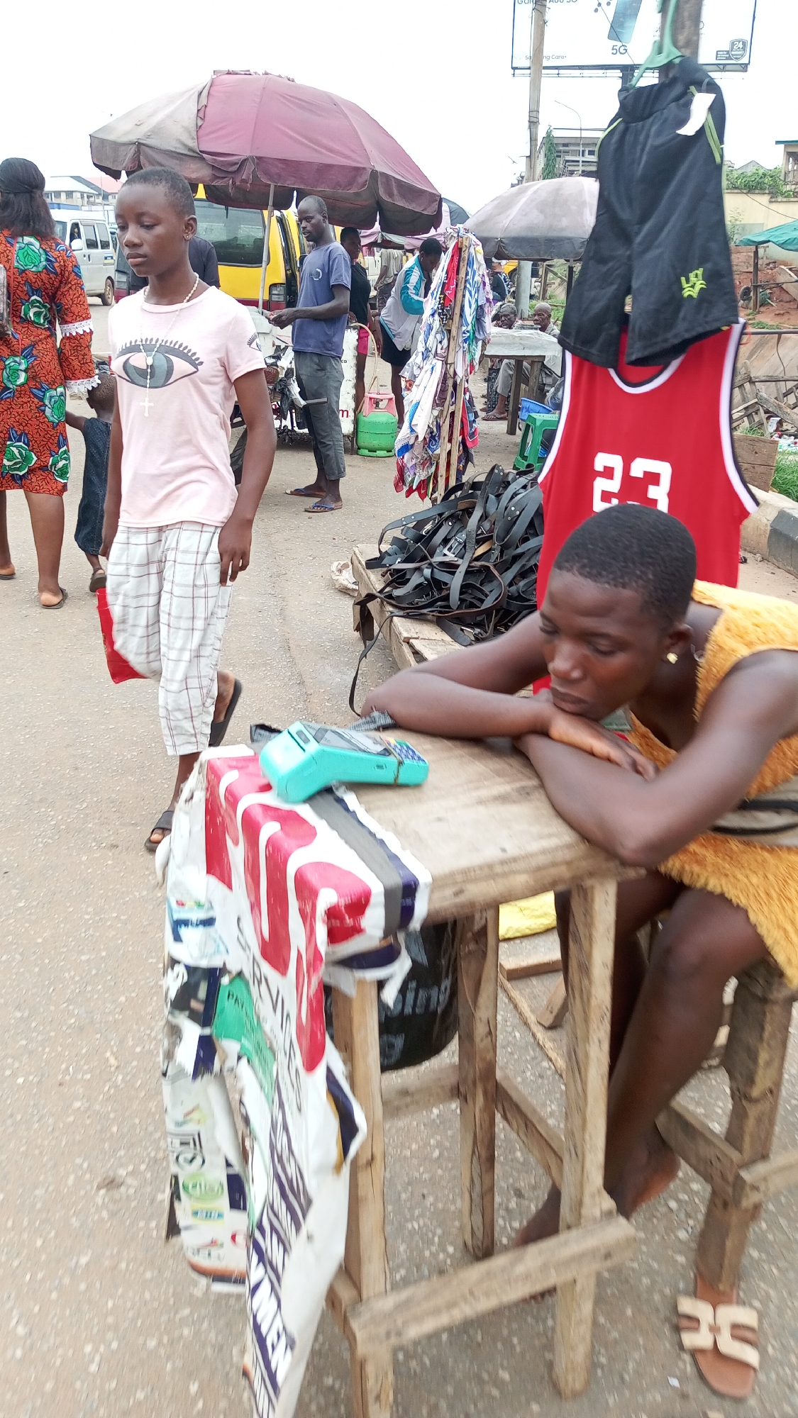 This image depicts a bustling street market scene in Onitsha, Nigeria, likely during the day under an overcast sky.
In the foreground on the right, a woman with short dark hair, wearing a yellow, textured top, leans her forearms on a rustic wooden table, resting her head on her hands. She appears contemplative or tired. Next to her on the table is a turquoise point-of-sale (POS) terminal. A printed banner, likely for advertisements or services, is draped over the front of her table, displaying partially visible words such as "SERVICES," "PAY," and "MEN." Her light brown sandals are visible near the table legs.
In the mid-ground on the left, a young girl with dark hair walks away from the viewer, looking forward with a focused expression. She wears a light pink t-shirt featuring a large eye design and a rosary-like necklace with a cross pendant, paired with white and red plaid trousers and black slide sandals. Behind her, a person in an orange patterned dress is partially visible.
Further into the mid-ground, under a large, faded red market umbrella, a man in a dark blue shirt and grey trousers stands near a yellow mini-bus. A green gas cylinder is visible near his stall. To the right of the umbrella, a clothing rack displays various fabrics and scarves. Suspended above a pile of black straps or belts are black athletic shorts with a green "LG" logo and a red basketball jersey with the number "23" on it.
In the background, more market stalls and umbrellas are visible, along with white and yellow vehicles. A billboard in the upper right background shows "5G" and other text related to a mobile carrier. The overall scene captures the vibrant daily commerce and activity typical of an urban market environment.